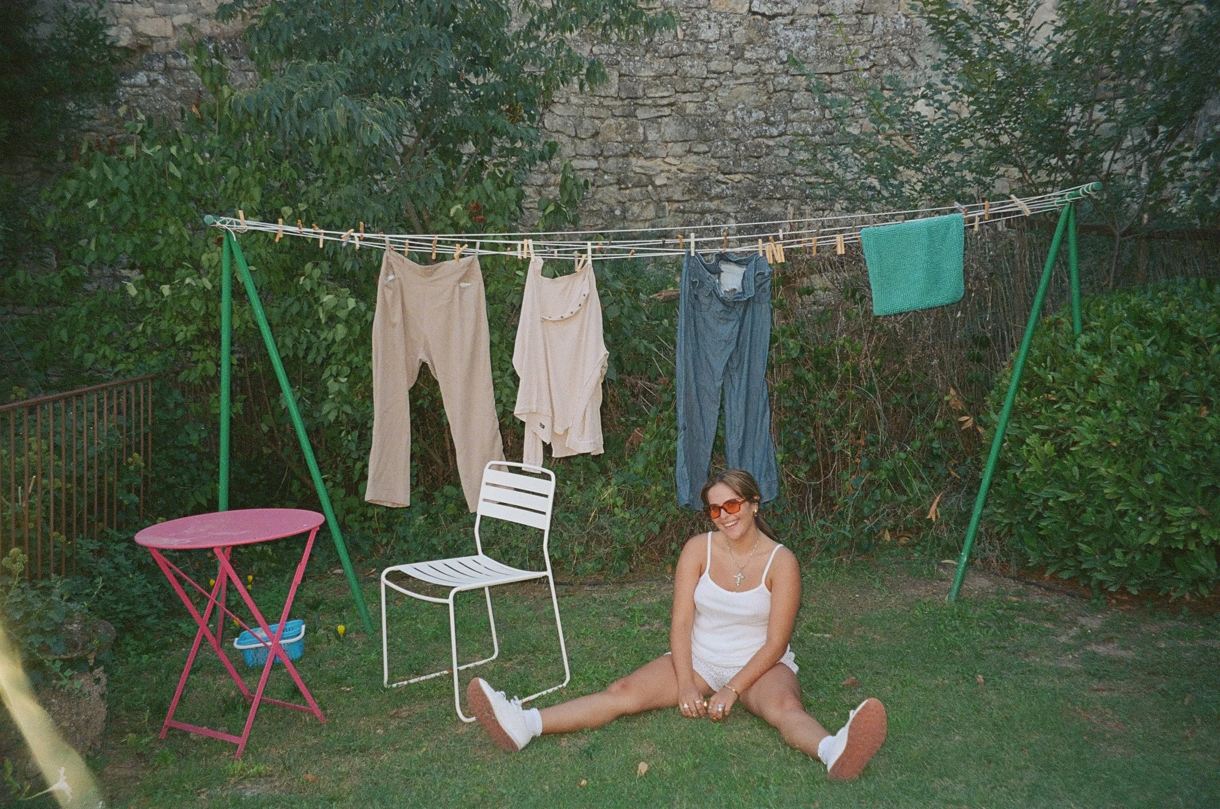 A young woman in white clothes and sunglasses sitting on the grass in a backyard with a clothesline hanging overhead. The clothesline has various clothing items hanging on it, and there are colorful outdoor chairs nearby. The background features a st