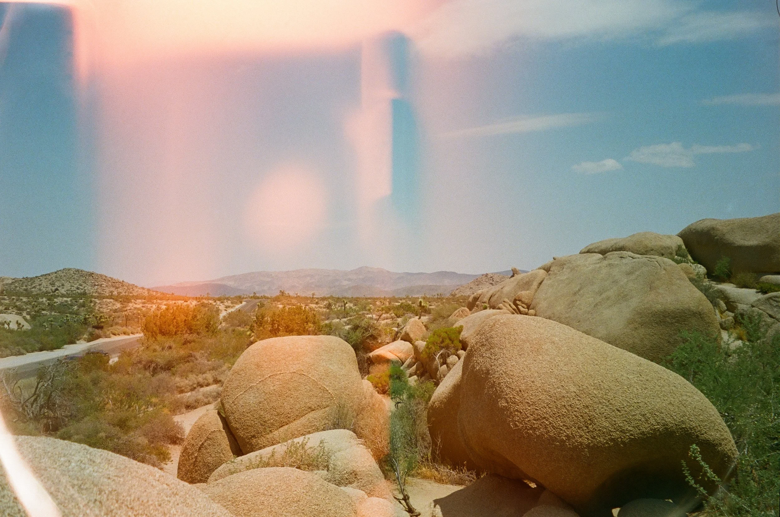 A desert landscape with large boulders in the foreground and mountain ranges in the background, under a partly cloudy sky with a faint rainbow