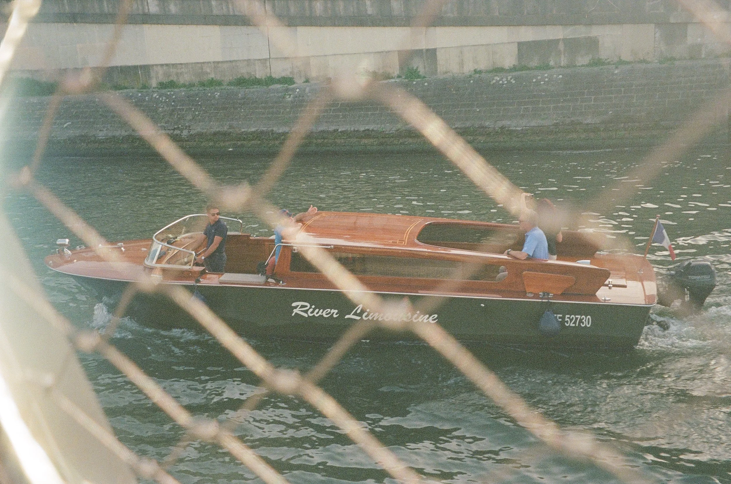 A wooden boat named 'River Limousine' is floating in a river with three people on board, two men and one woman. The boat is seen through a chain-link fence, and a concrete wall is visible in the background.