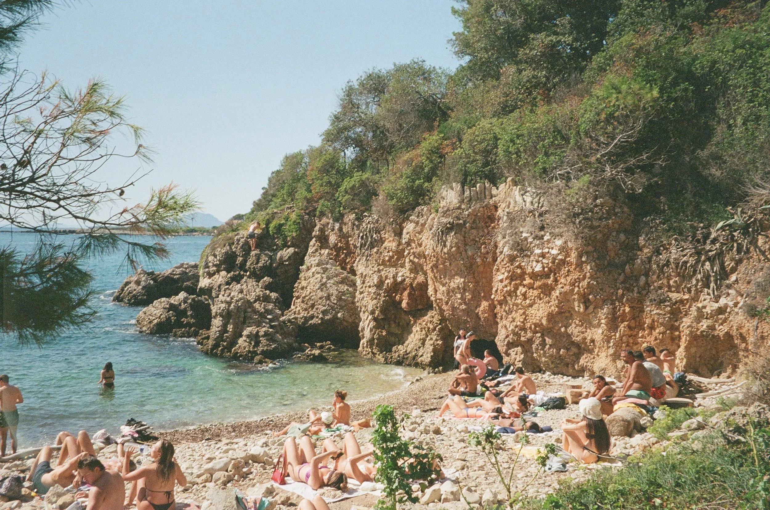 Beach scene with people relaxing and swimming by rocky cliffs and greenery, with trees and the ocean in the background.