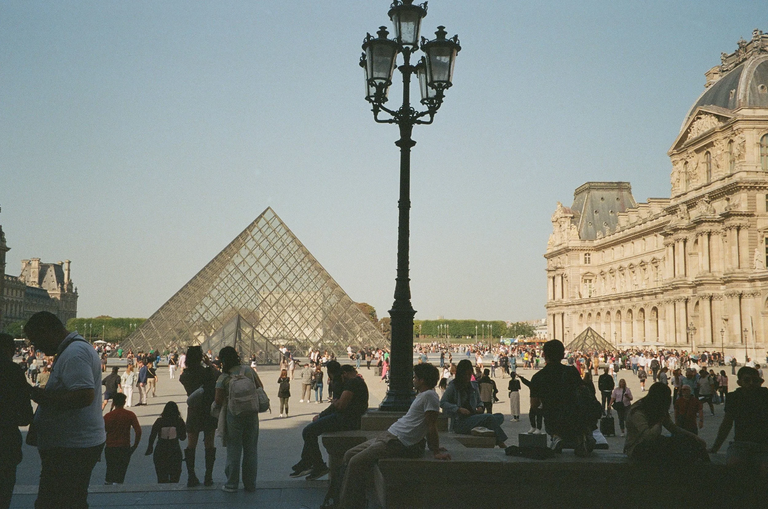 Crowd of people gathered in front of the Louvre Pyramid in Paris, France, with historic buildings in the background.