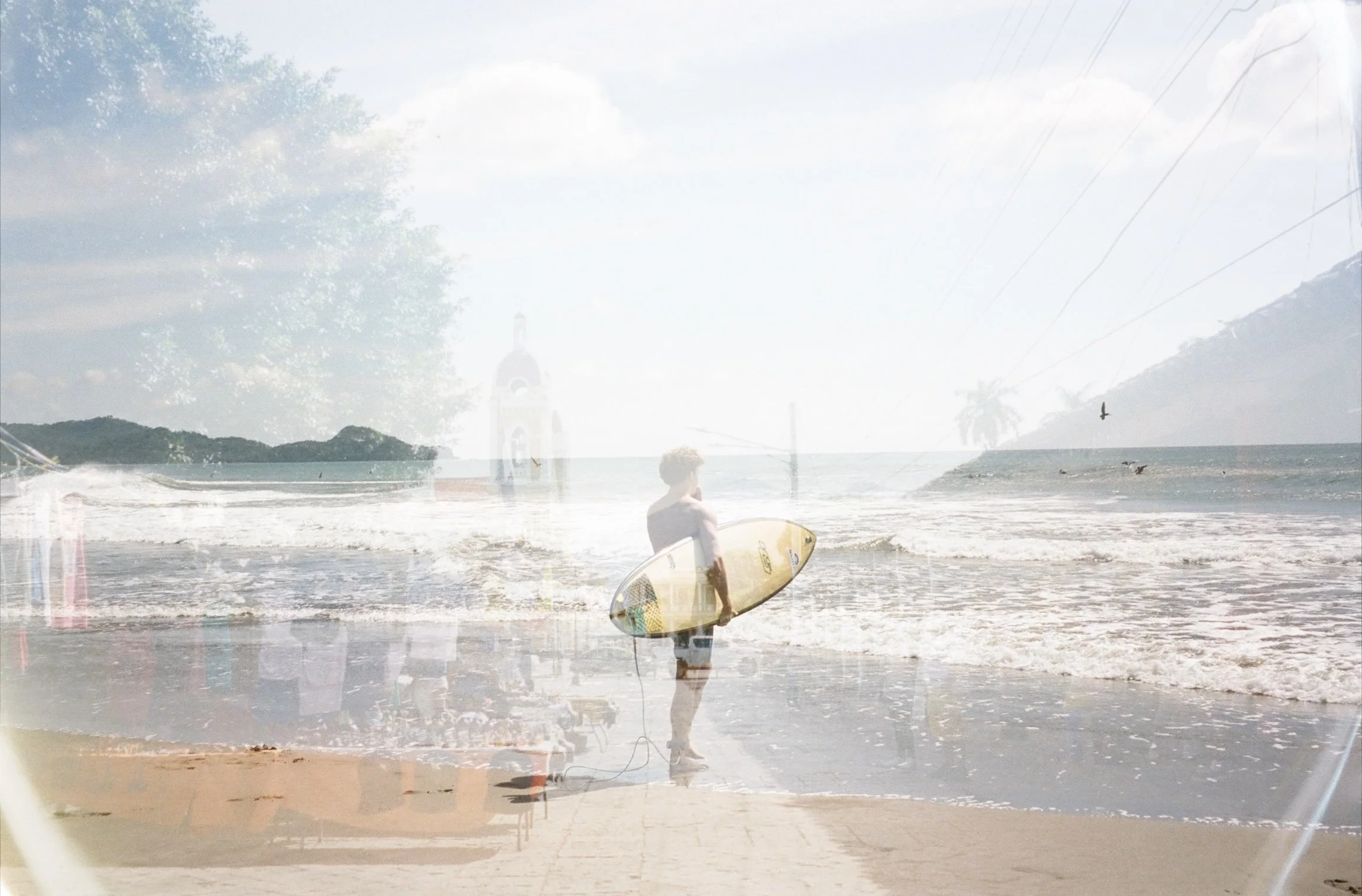 A person standing on the beach holding a surfboard, with a view of the ocean, mountains, and palm trees in the background, under a partly cloudy sky.