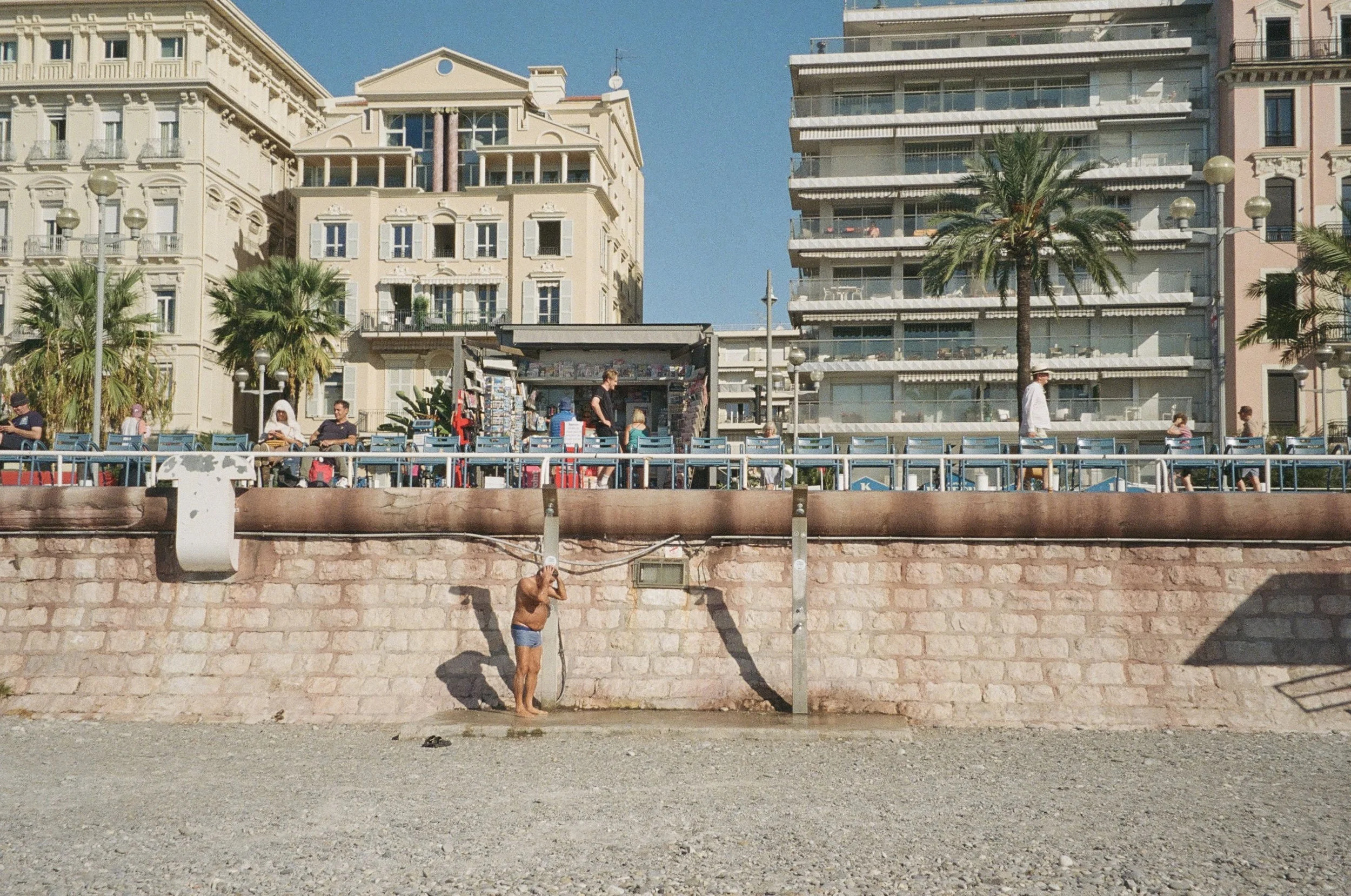 A man stands on a beach with his hand raised to his ear, next to a stone wall beneath a promenade with people sitting and walking, backed by tall buildings and palm trees under a clear blue sky.