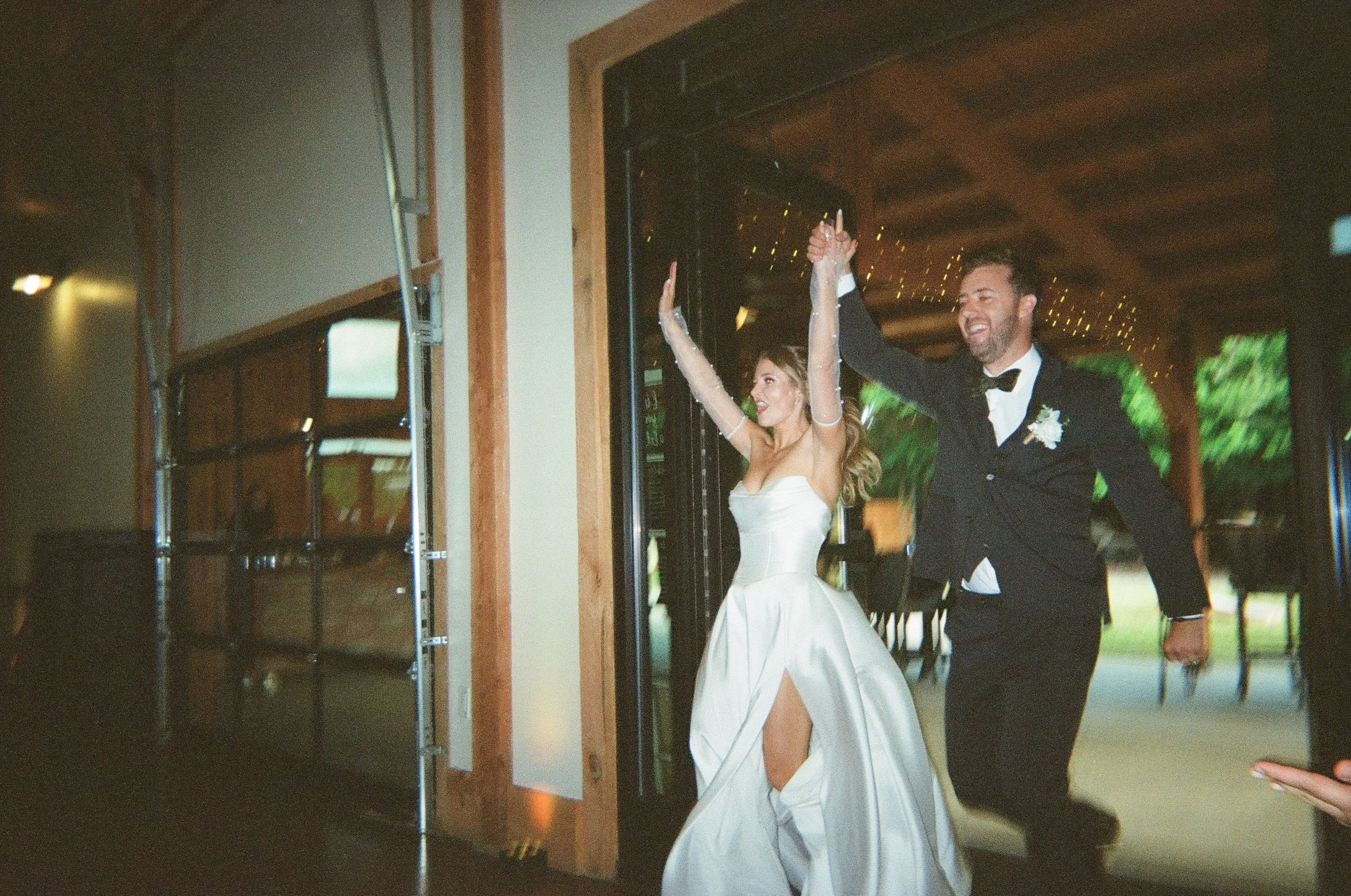 A bride and groom celebrating at a wedding reception, holding hands and smiling, with the bride in a strapless white dress and the groom in a black tuxedo.