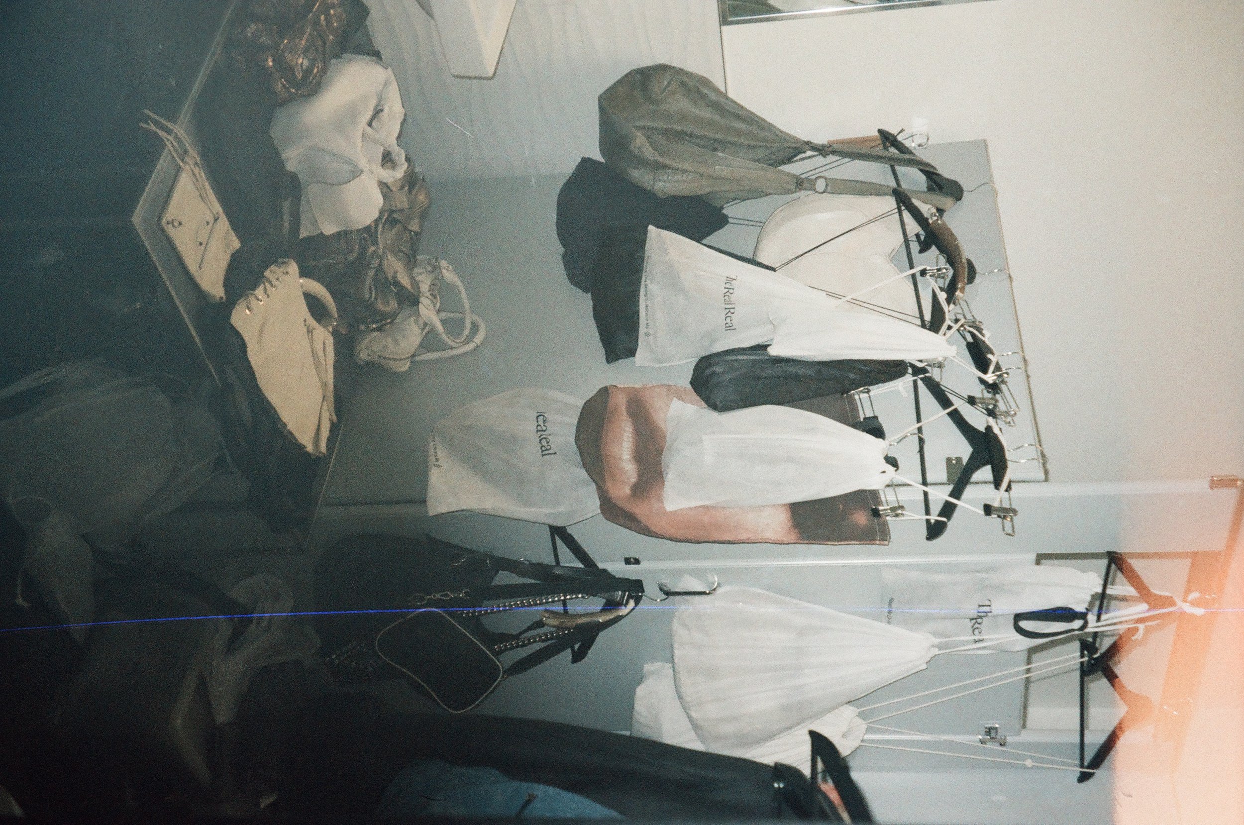 Clothes hanging on a clothes rack, including shirts and jackets, with shopping bags and shoes nearby in a retail or storage space.