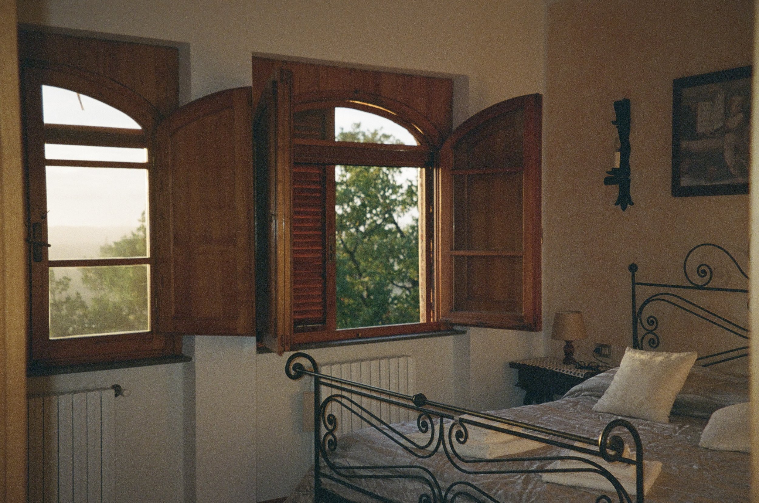 A bedroom with a wrought iron bed, a lamp, and a window with wooden shutters open, showing trees outside.