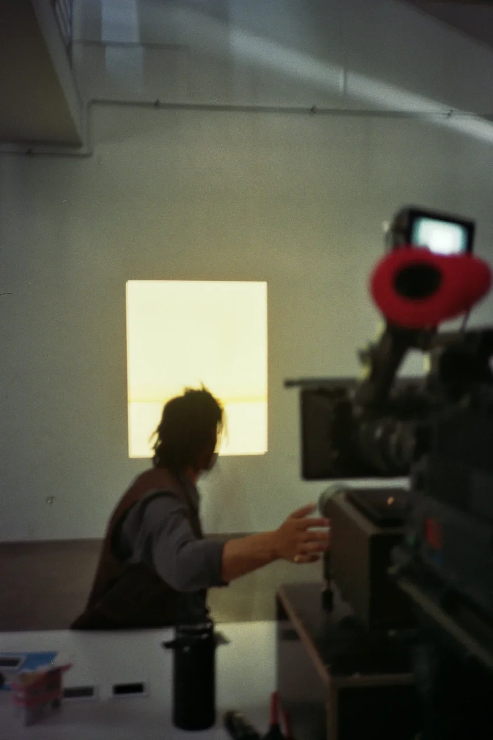 A person with dreadlocks interacting with equipment, seen from behind, with a large camera in the foreground capturing the scene. There is a bright square light on the wall behind them.