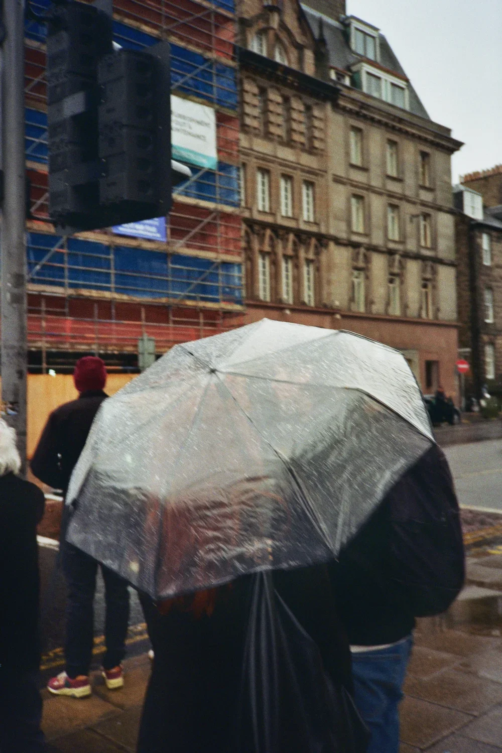 People standing on a city street in rainy weather, with some holding umbrellas, and a building under construction in the background.