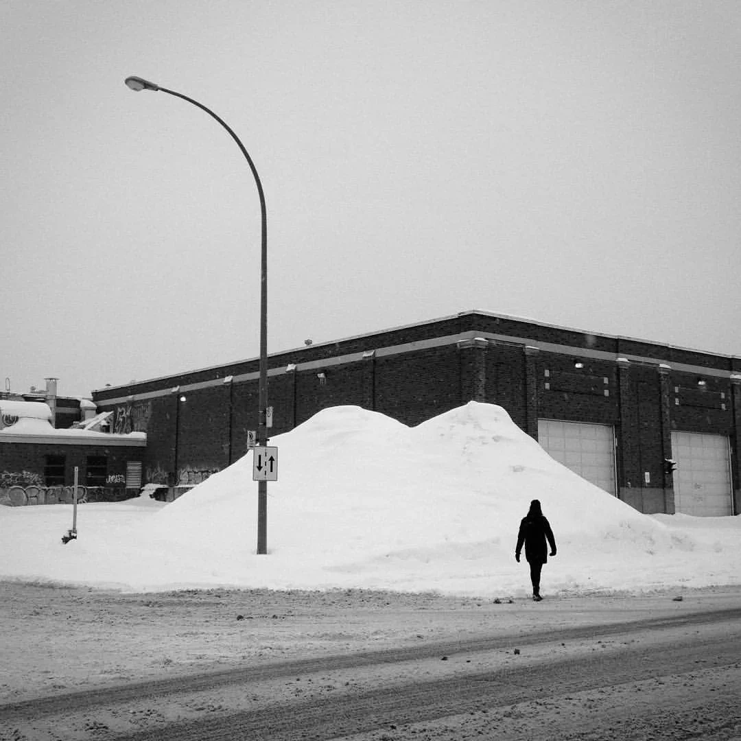 A person walking on a snowy street near a large pile of snow in front of a brick building, with a streetlamp and traffic sign visible.