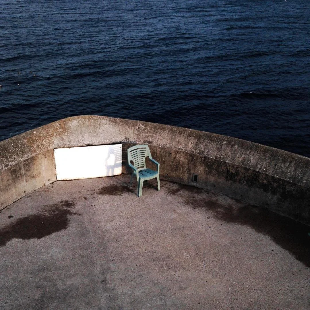 A single plastic chair on a concrete deck next to a curved stone railing overlooking the ocean at dusk.