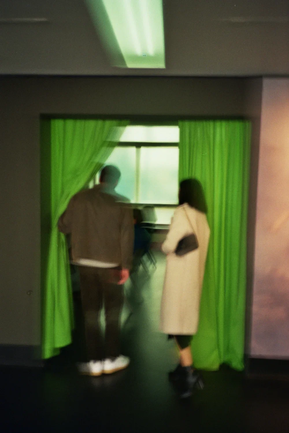 Two people, a man and a woman, stand near green curtains and look out of a window.