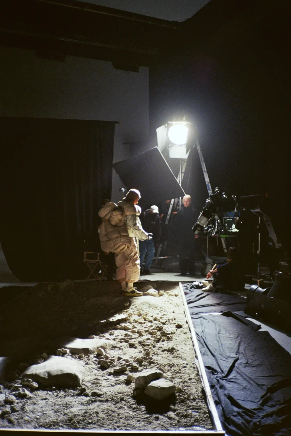 Behind the scenes of a film set, crew members prepare for filming on a sandy surface with a black curtain backdrop, bright lighting, and camera equipment.