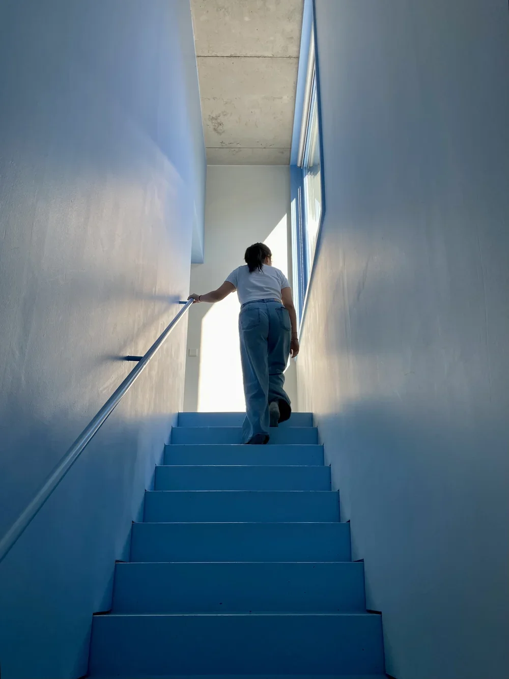 Person ascending a bright staircase in a modern building with blue walls and large window letting in natural light.