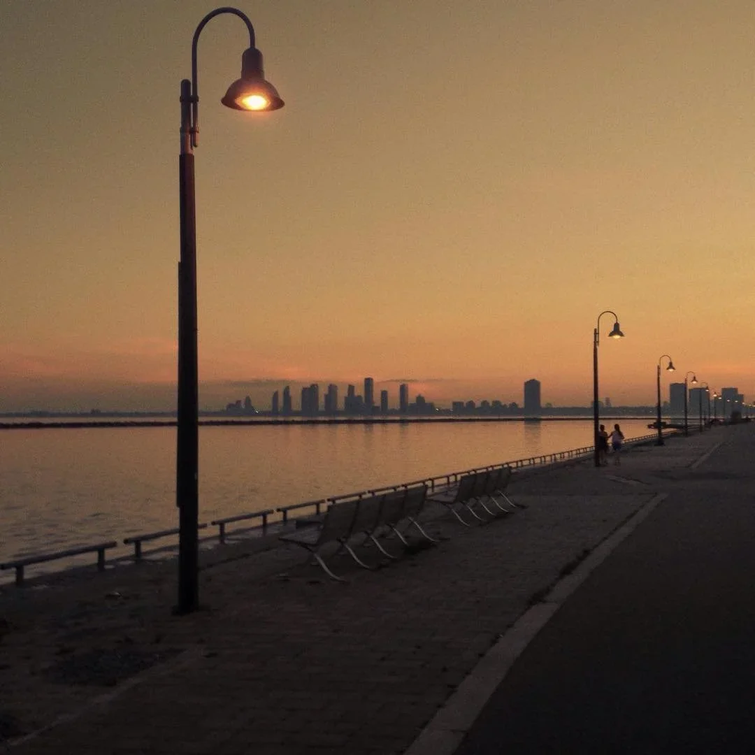 A waterfront promenade at sunset with street lamps lit, benches, and a city skyline in the distance.