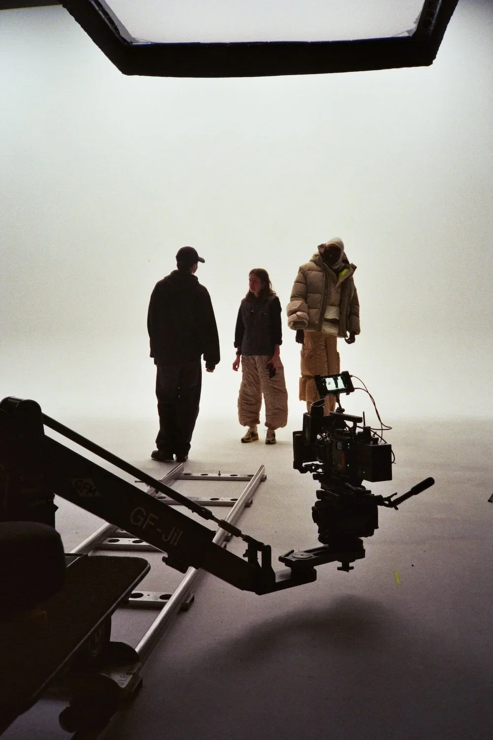 Three people standing on a white seamless background during a filming or photoshoot. Equipment and a camera crane are visible in the foreground.