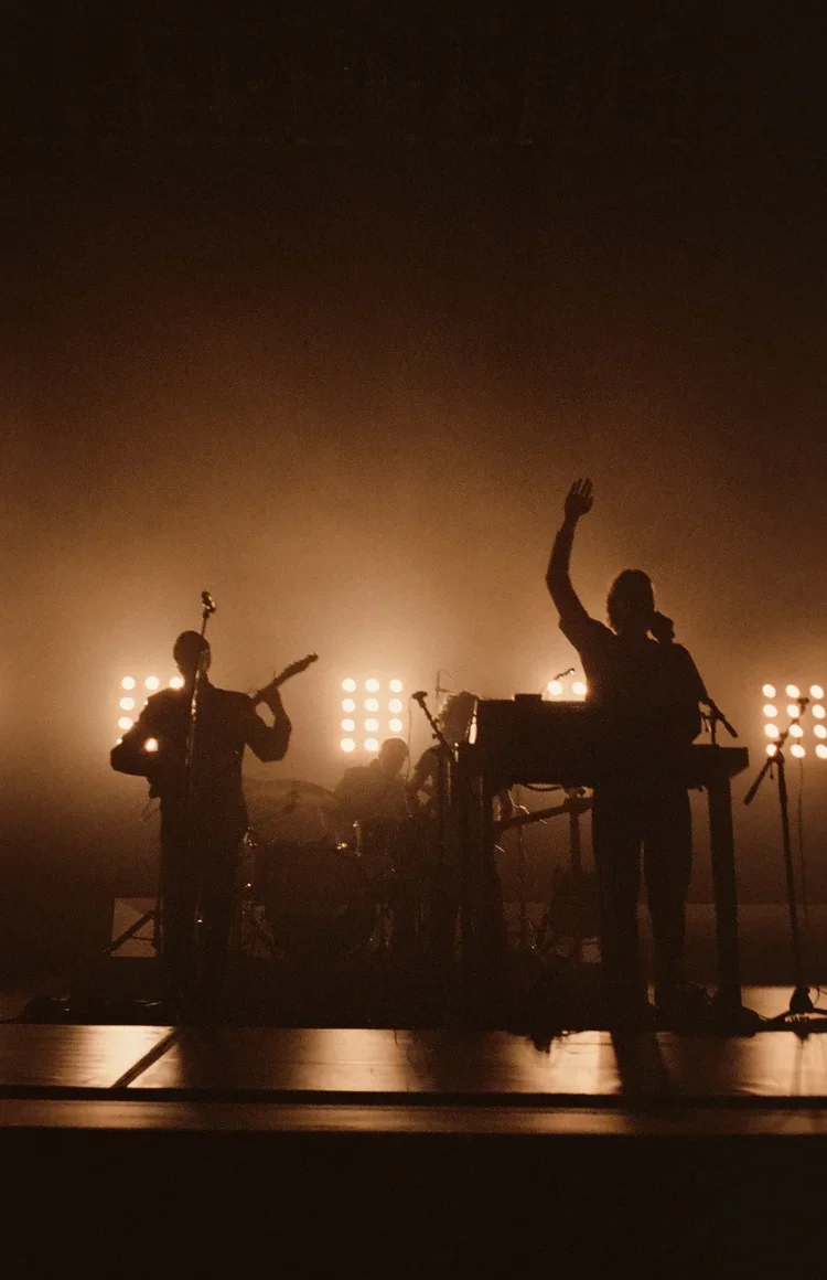 Silhouettes of a band performing on stage with bright lights behind them, including a guitarist, a keyboardist, and a drummer.