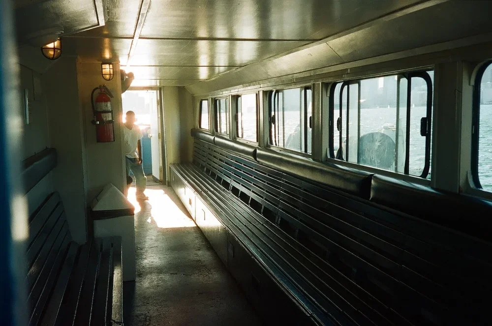 Empty ferry boat interior with black benches, windows showing water outside, and a person near the door in the background.