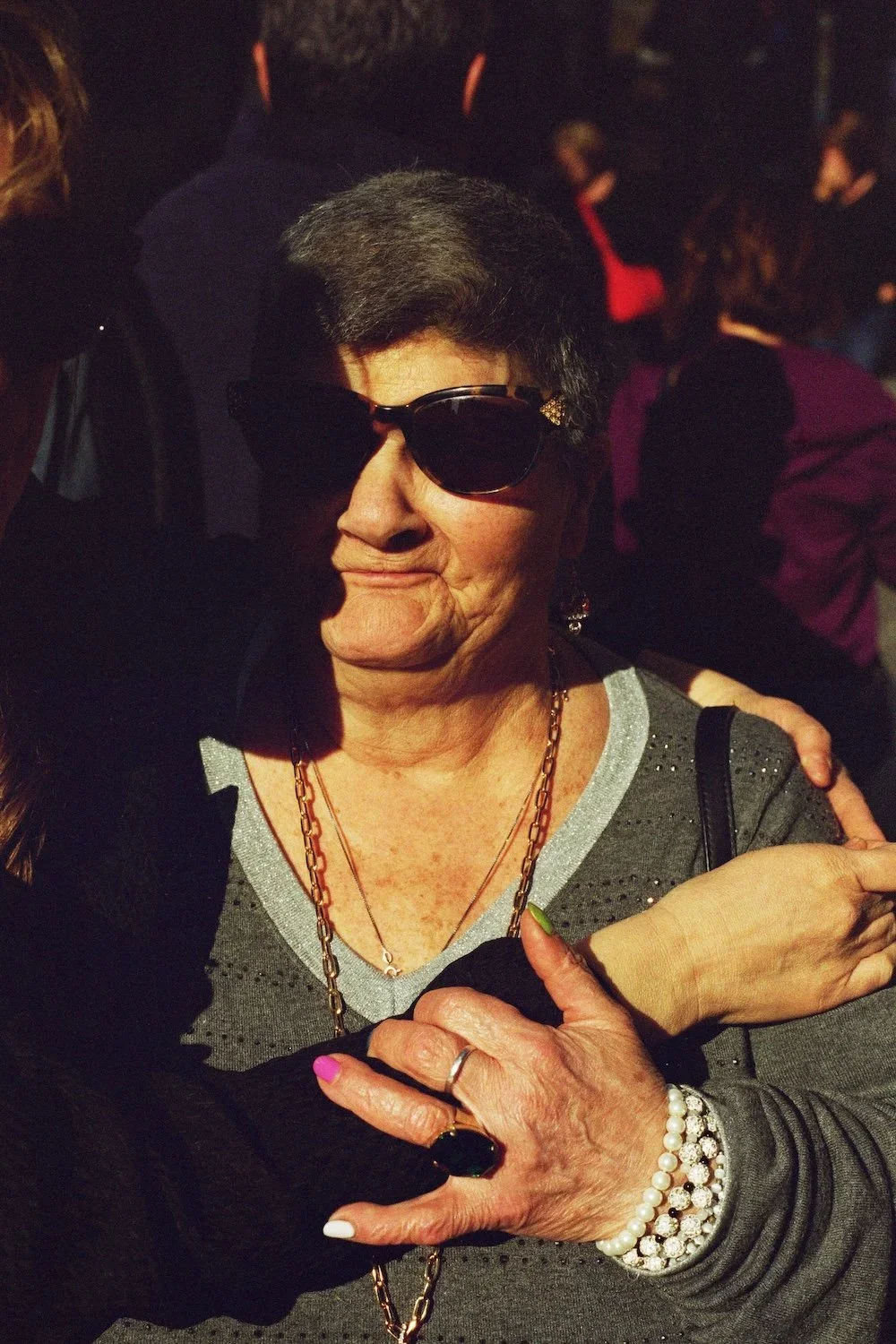 An older woman wearing sunglasses and a gray top, exchanging an embrace with another person. She has jewelry including a pearl bracelet, rings, and layered necklaces.