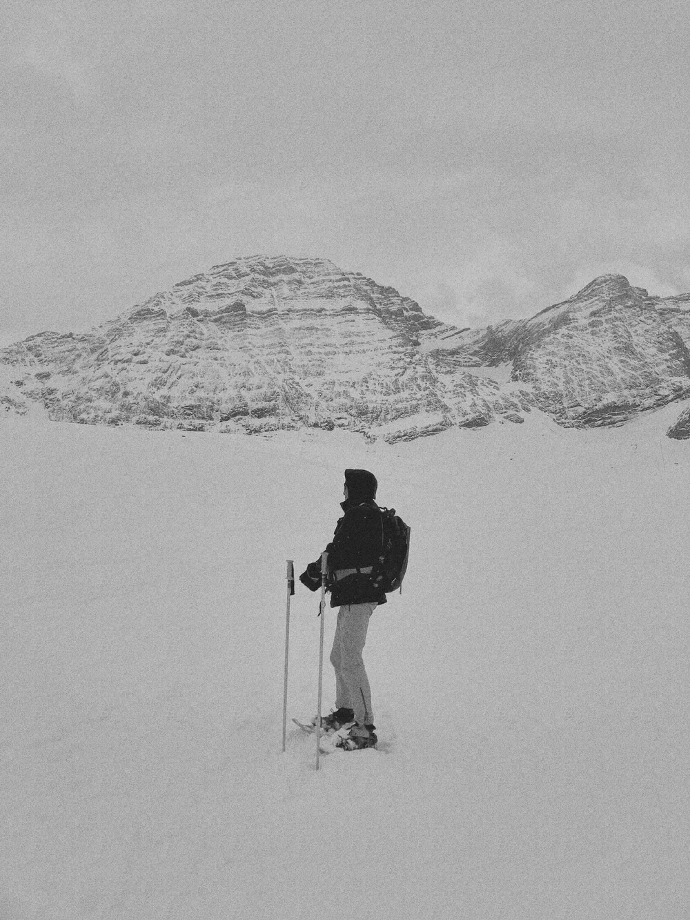 A person dressed in winter gear stands in snowy terrain with mountains in the background.