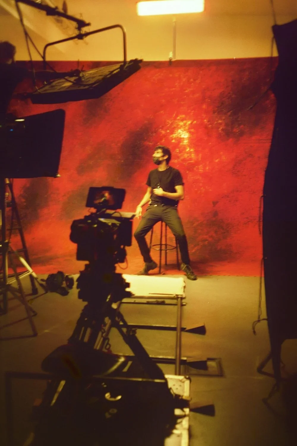 Person standing on a stool in front of a red textured backdrop, holding a microphone during a photoshoot or filming session with camera and lighting equipment around.