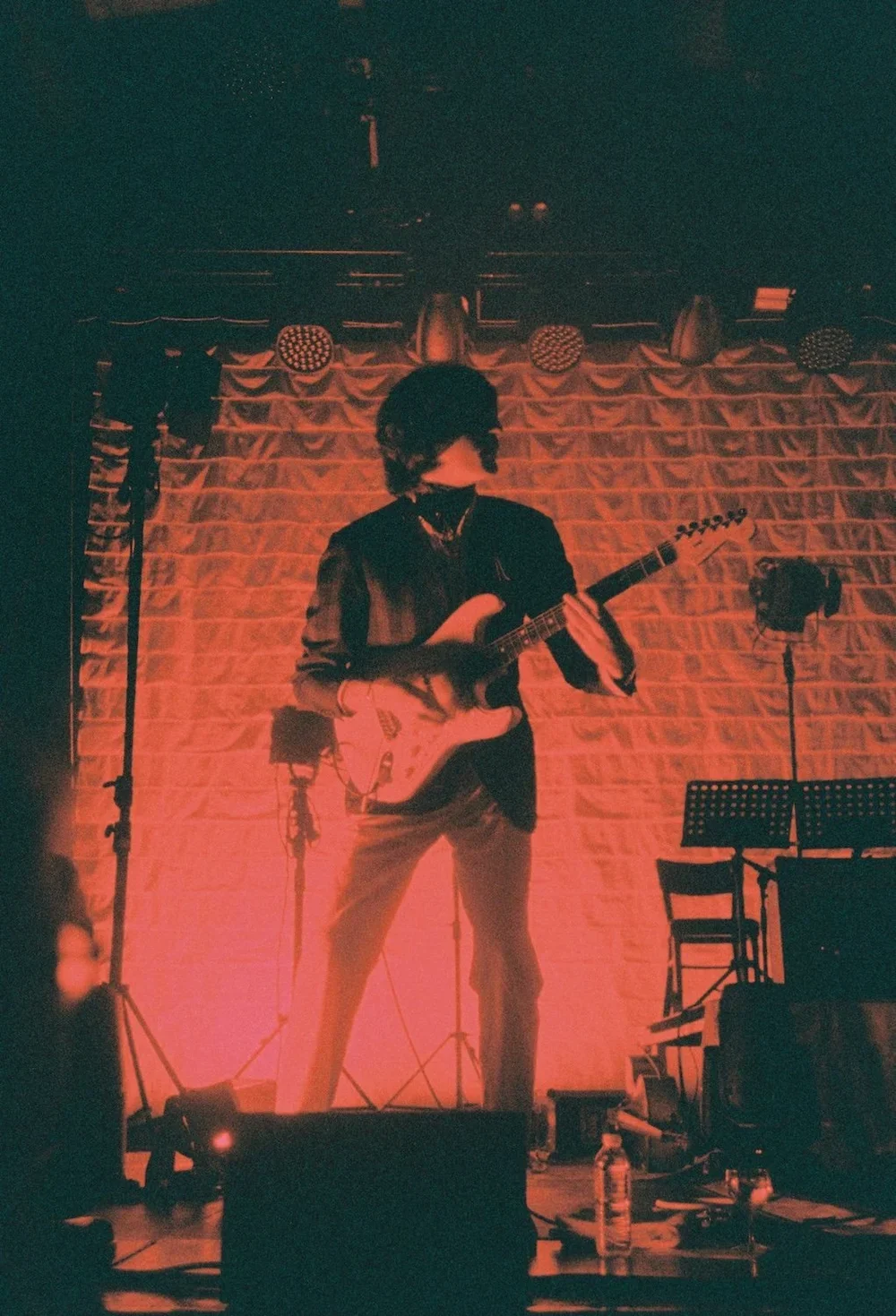 A musician playing an electric guitar on stage with a brick wall backdrop, stage lights, and music stands.