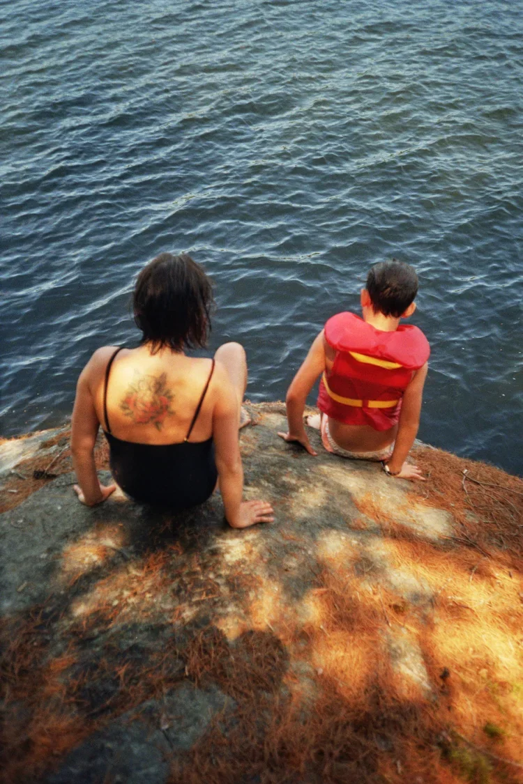 A woman and a boy sitting on a rocky ledge by the water, with the woman wearing a black swimsuit and the boy with a red life jacket, looking out over the water.