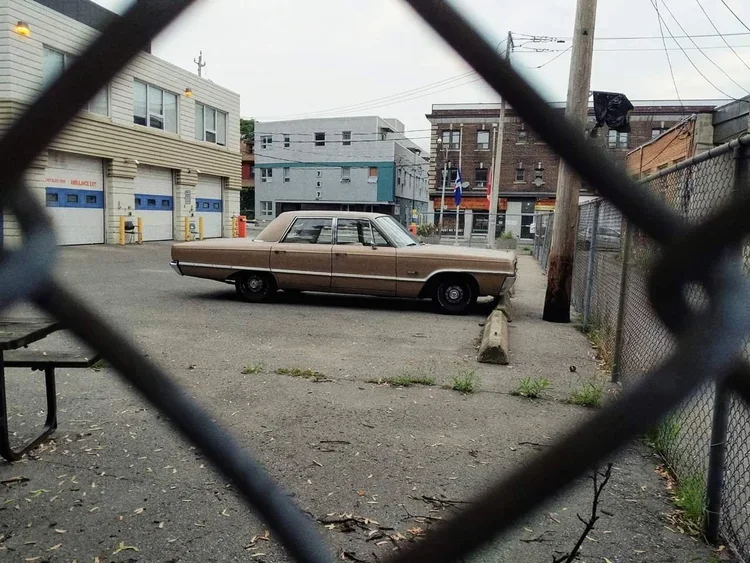 An old, beige car parked behind a chain-link fence in an urban alley, with buildings, utility poles, and a chain-link fence visible in the background.