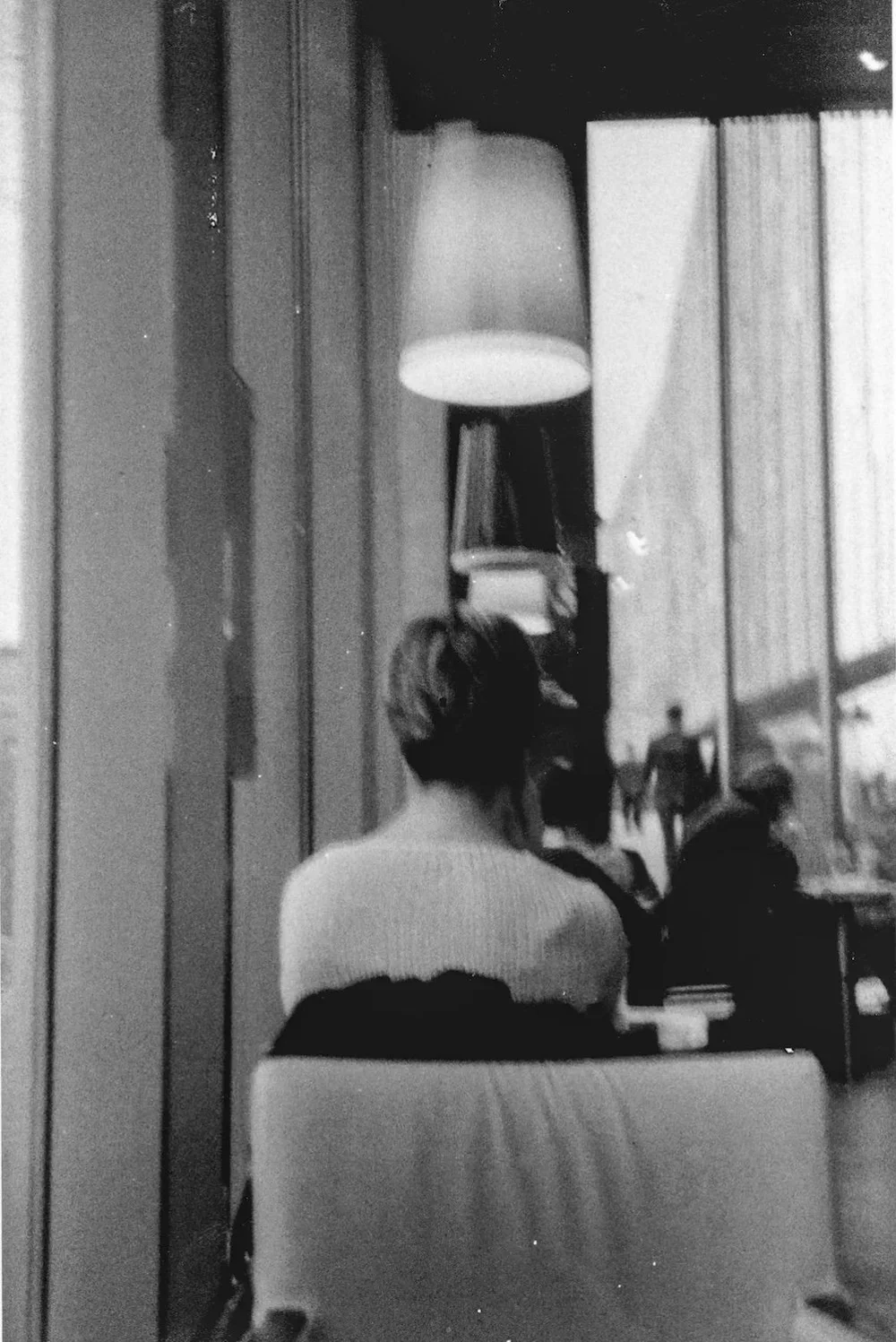 A person with short hair sitting in a cafe, viewed from behind, looking out the window.