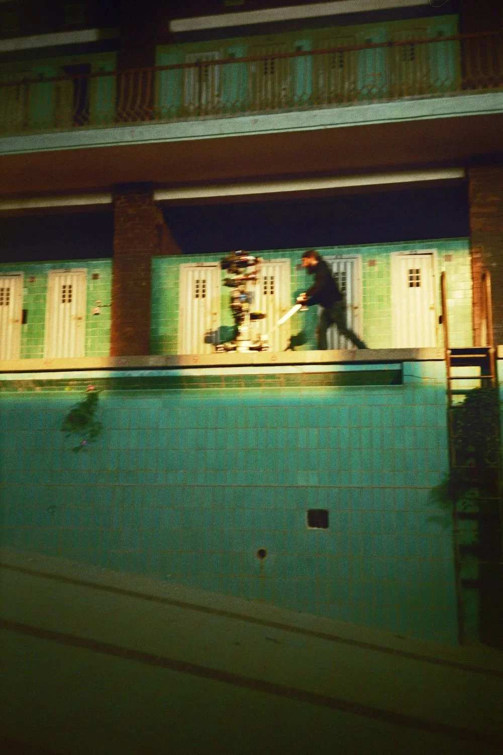 A person cleaning or maintaining a tiled wall at the edge of an empty swimming pool at night, illuminated by artificial lighting.