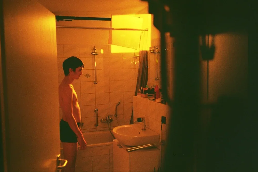 A young man standing in a dimly lit bathroom, looking down, with a bathtub and sink visible in the background.