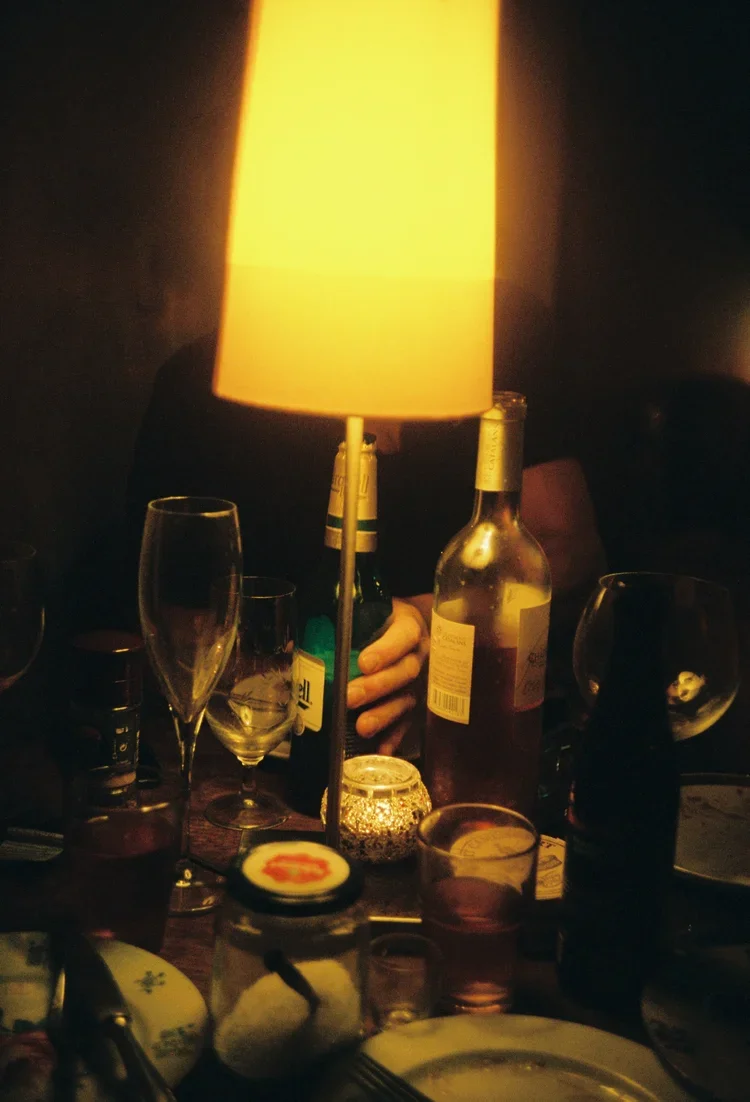 A dimly lit table with a yellow lampshade, various wine glasses, beer bottles, a candle holder, and some plates and utensils.