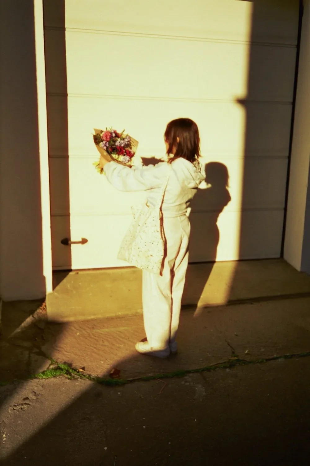 A person holding a bouquet of flowers, standing in the sunlight in front of a garage door, casting a shadow.