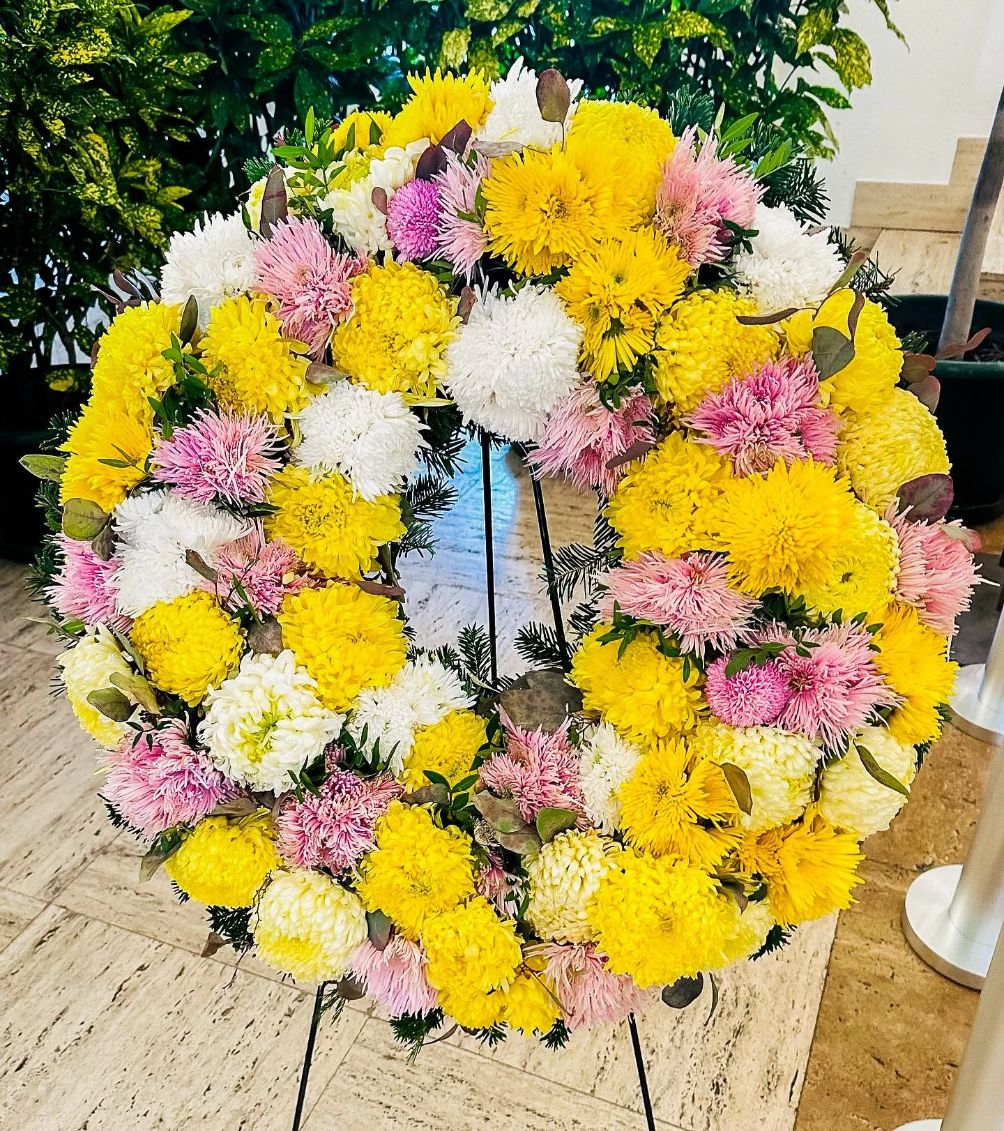 Blumenkranz mit gelben, weißen und rosa Chrysanthemen auf einem schwarzen Metallständer.