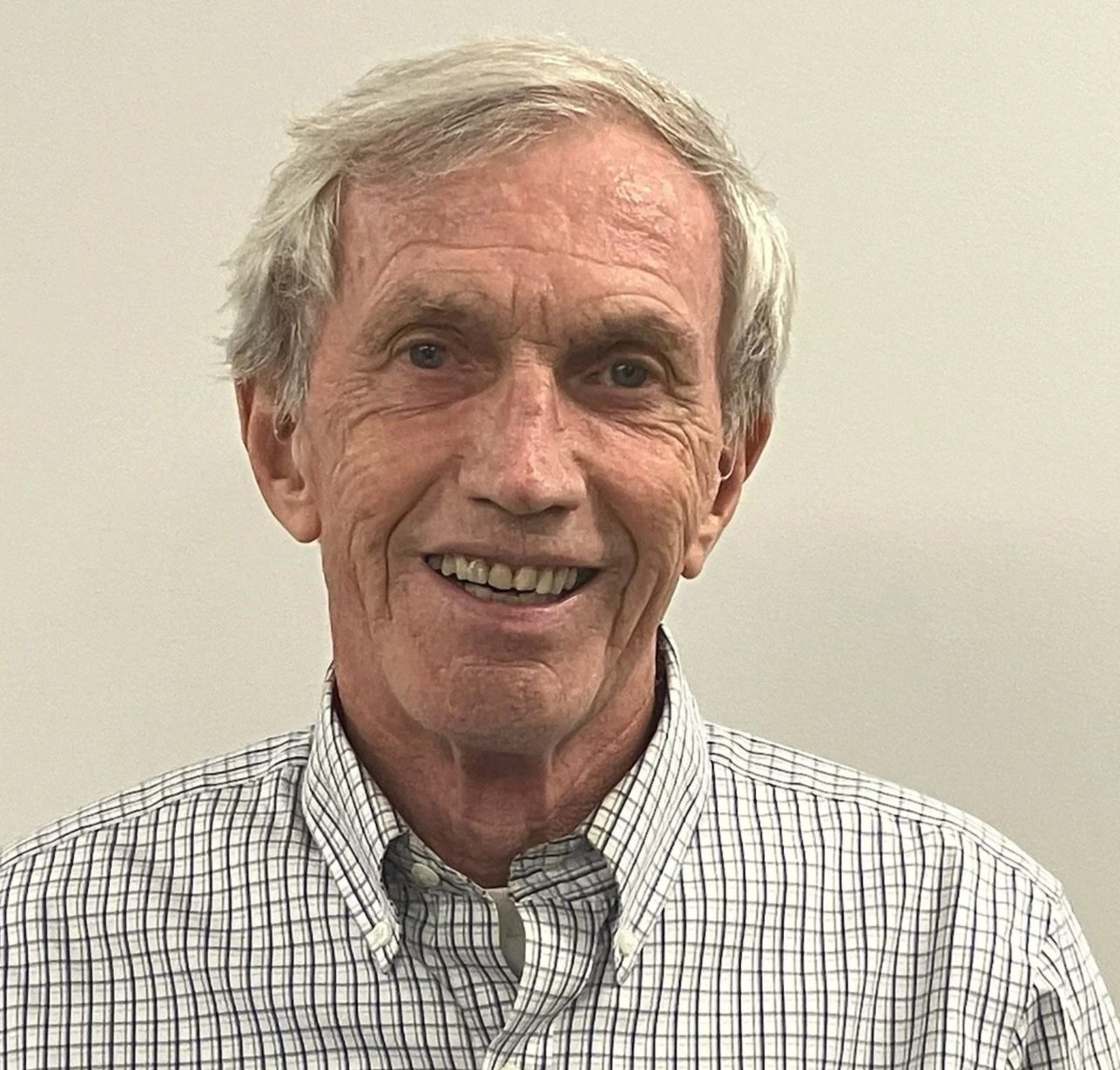 A smiling older man with gray hair, wearing a checkered shirt, standing against a plain light-colored background.