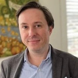 A man with dark brown hair, wearing a gray blazer and light blue shirt, smiling indoors with a window and blurred greenery in the background.