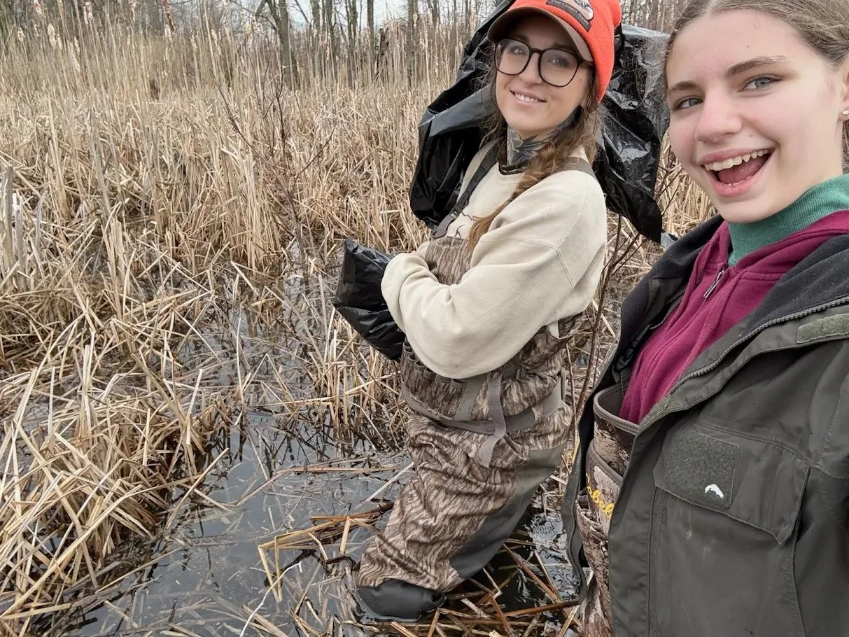 Two young women outdoors in a marshy area with tall brown grass, smiling at the camera during a hike or outdoor activity.