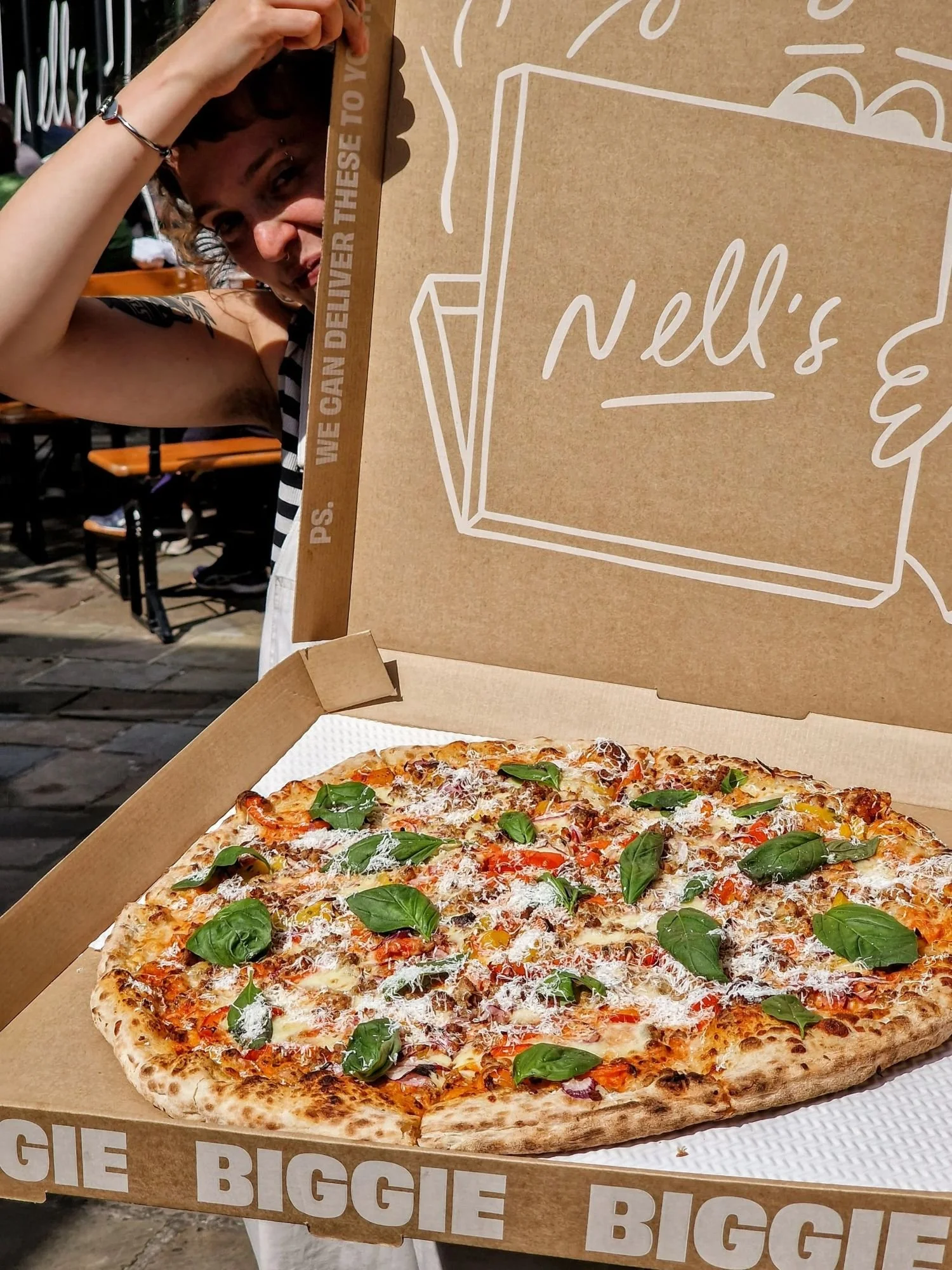A freshly baked pizza topped with basil leaves, cheese, and vegetables in an open pizza box with "Nell's" written on the inside of the lid.