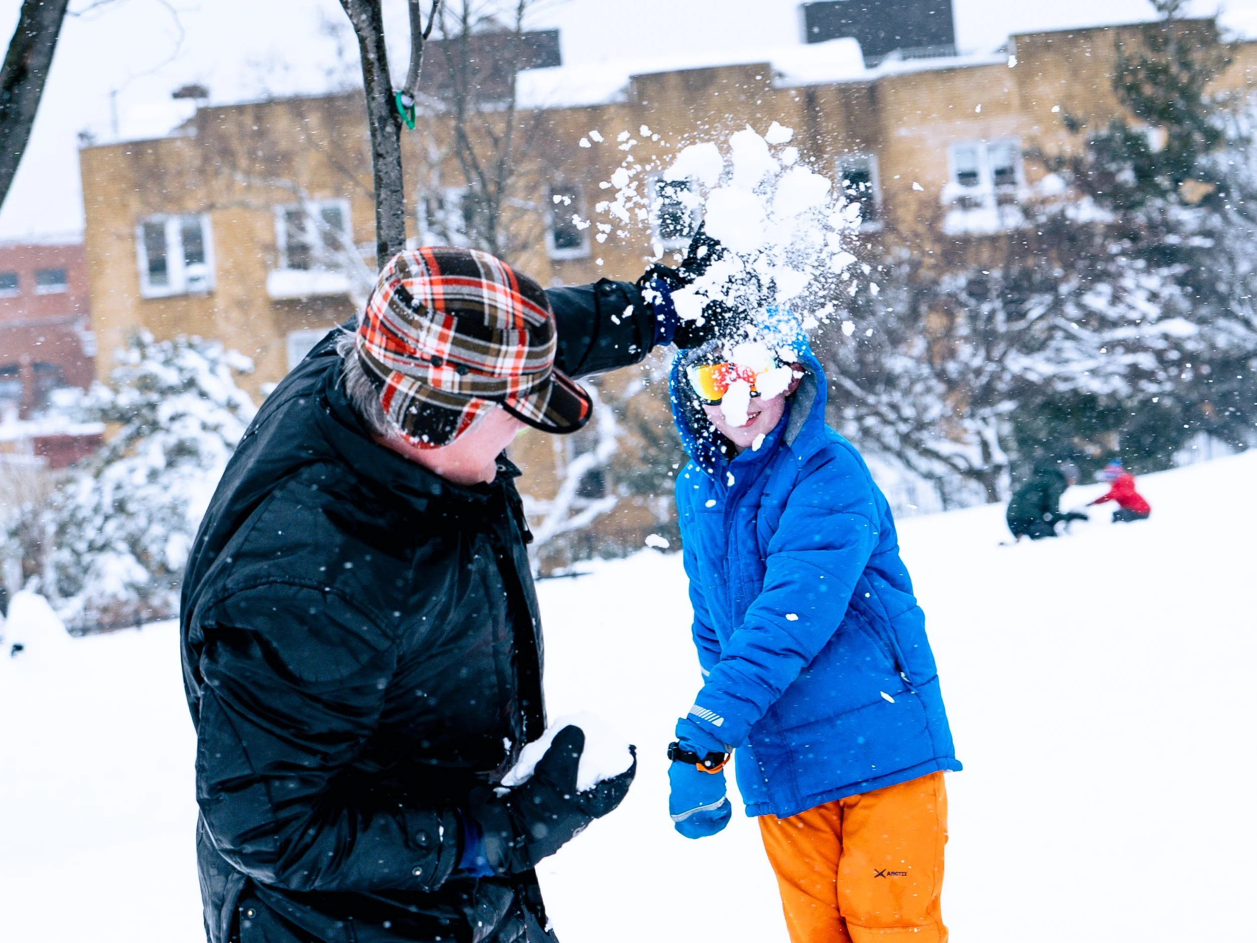 A man and a child playing in the snow, with the man hitting the child with a snowball, both dressed warmly in winter clothing, with snow-covered trees and buildings in the background.