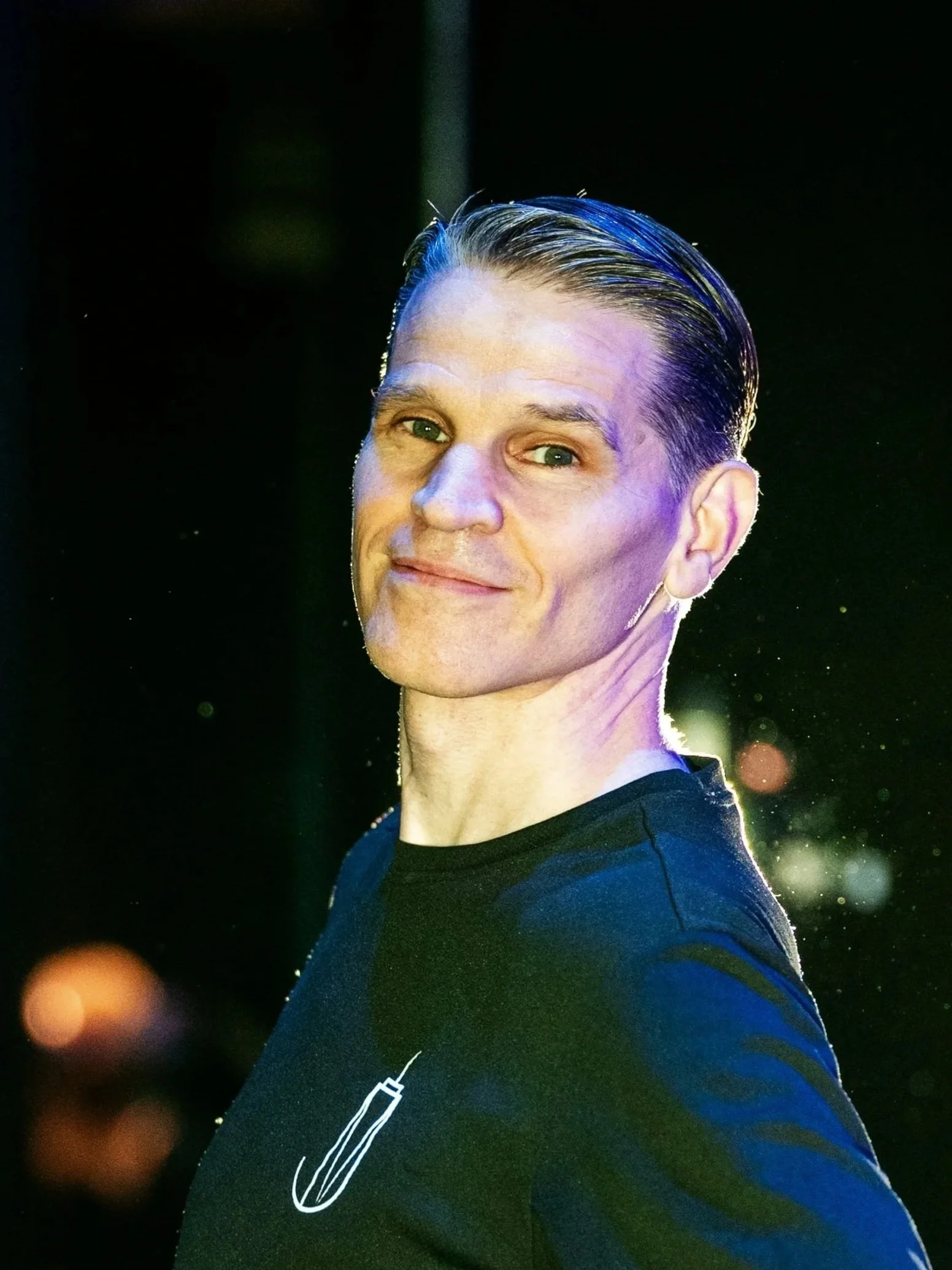 A man with slicked-back hair wearing a black t-shirt with a small white design near his collar, standing with one hand on his hip and smiling at the camera, illuminated by colorful lighting in a dark environment.
