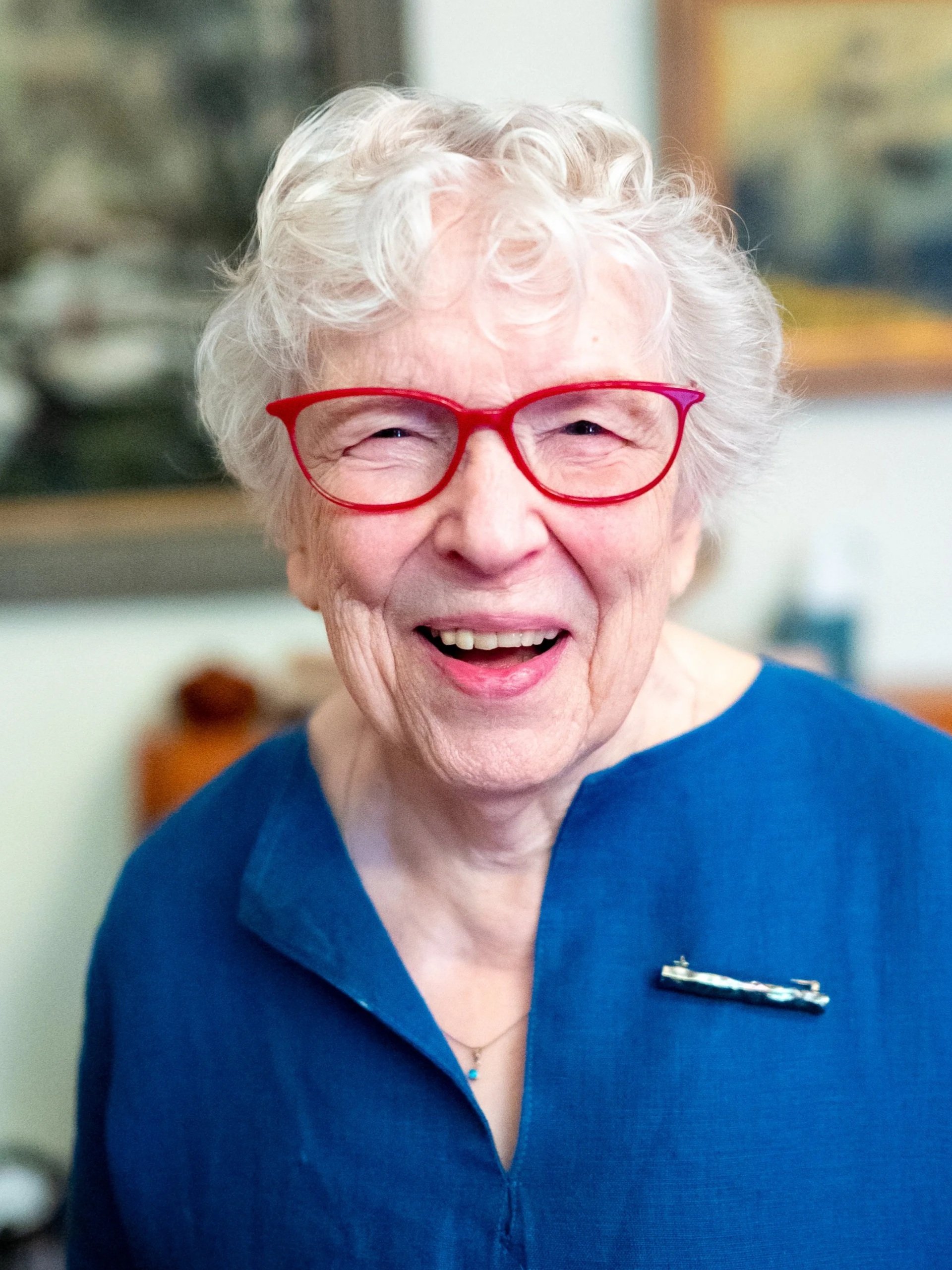 An elderly woman with white curly hair wearing red glasses and a blue top, smiling in a room with paintings on the wall.