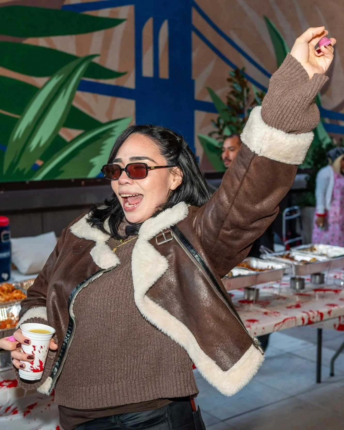 A woman with black hair, wearing sunglasses and a brown shearling jacket, is smiling and raising her arm while holding a small cup in her left hand. She appears to be at a festive gathering with food trays on a table in the background.