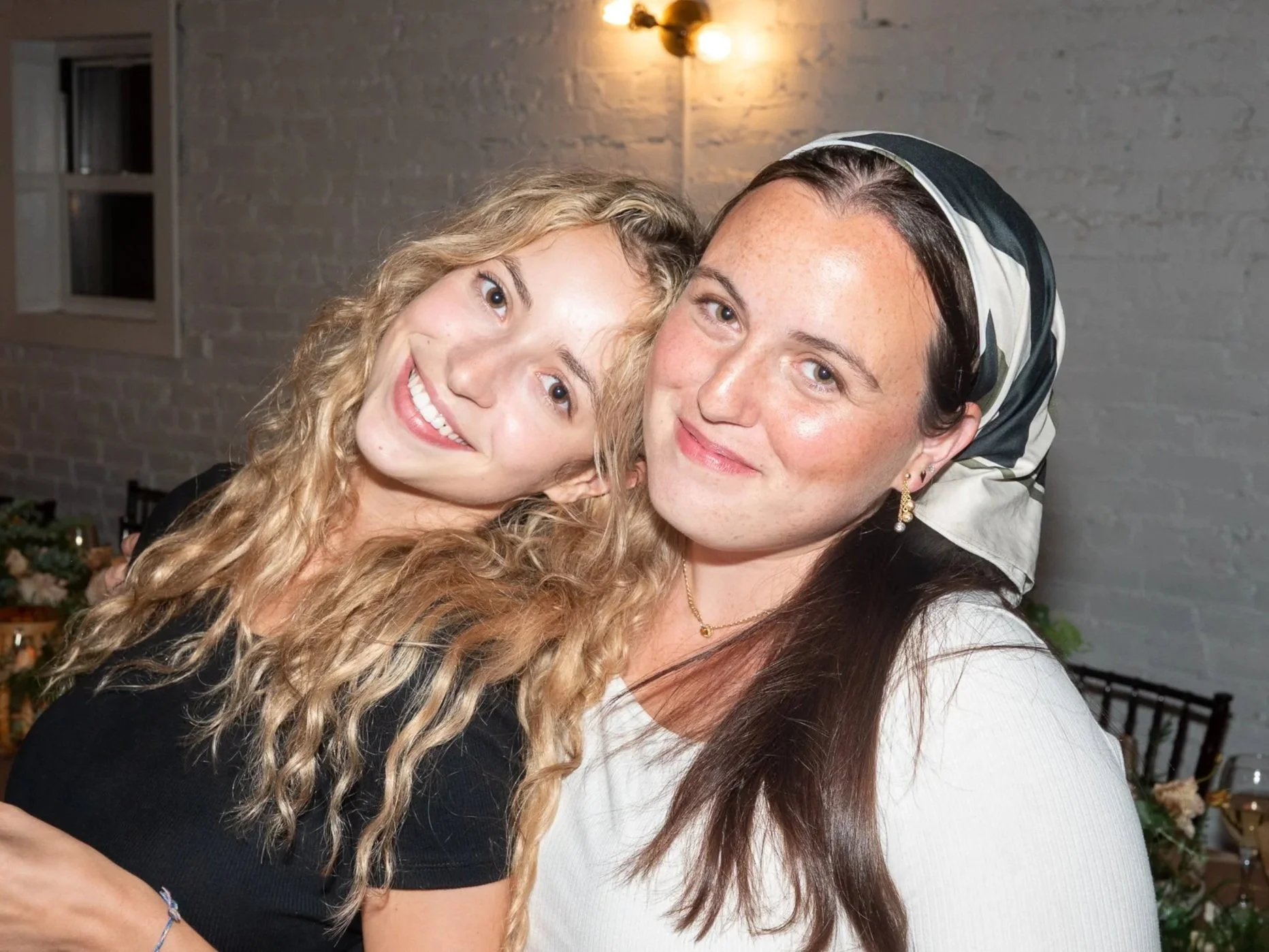 Two women smiling and embracing at a dinner party, with decorated tables and white brick walls in the background.