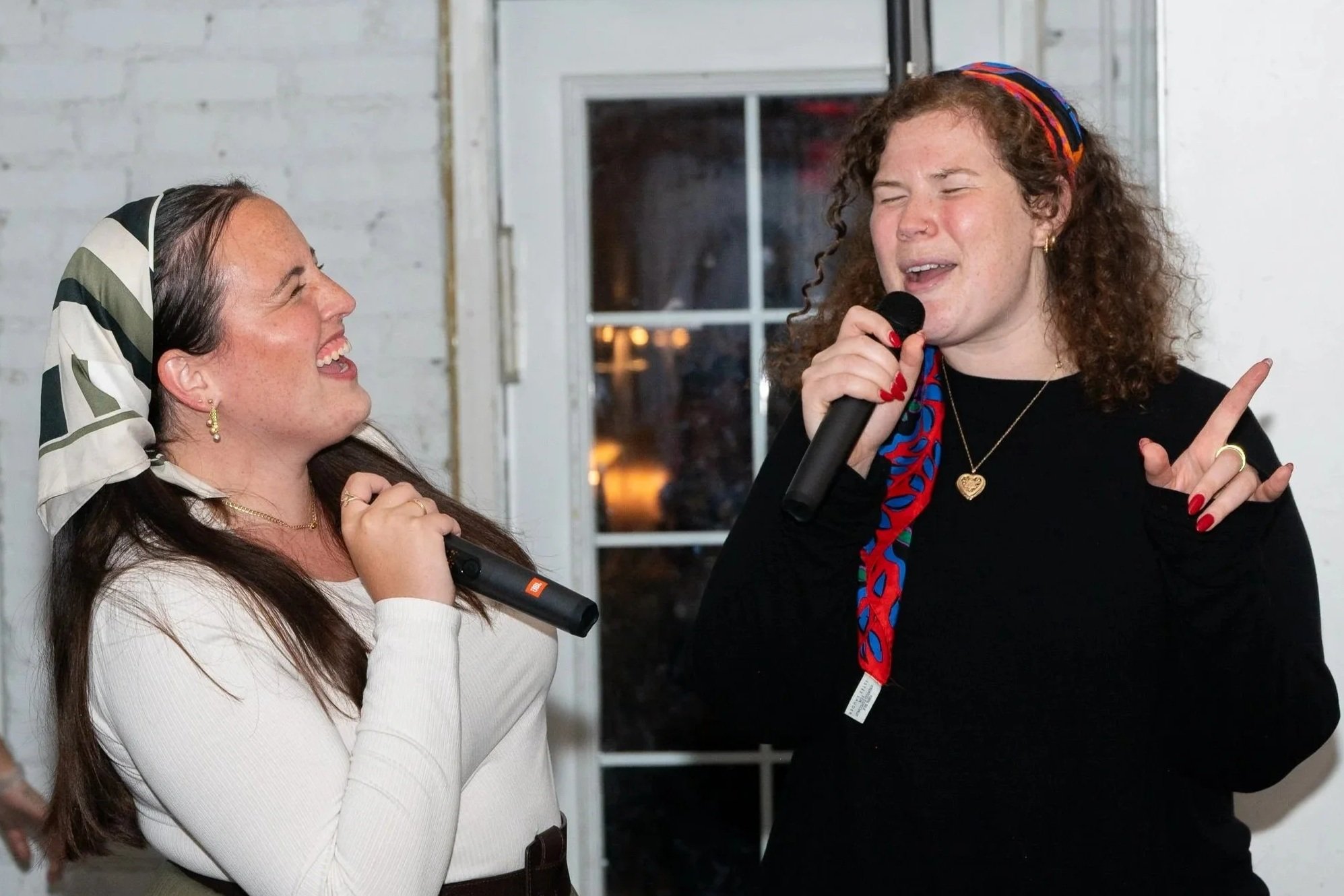 Two women singing karaoke together, smiling and enjoying the moment indoors with a white brick wall and window in the background.