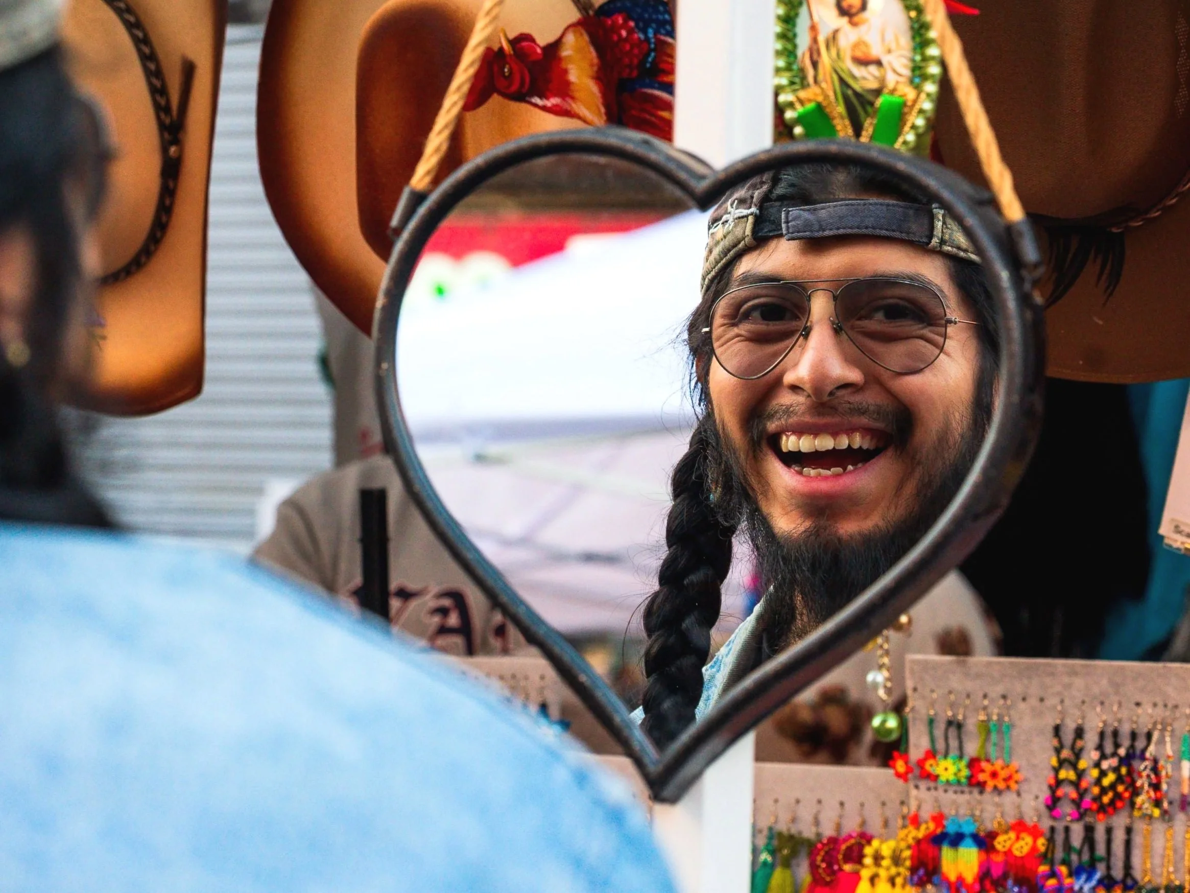 A man with long hair, braided beard, and glasses looks into a heart-shaped mirror, smiling. The mirror is surrounded by colorful jewelry and decorations, with other people visible around him.