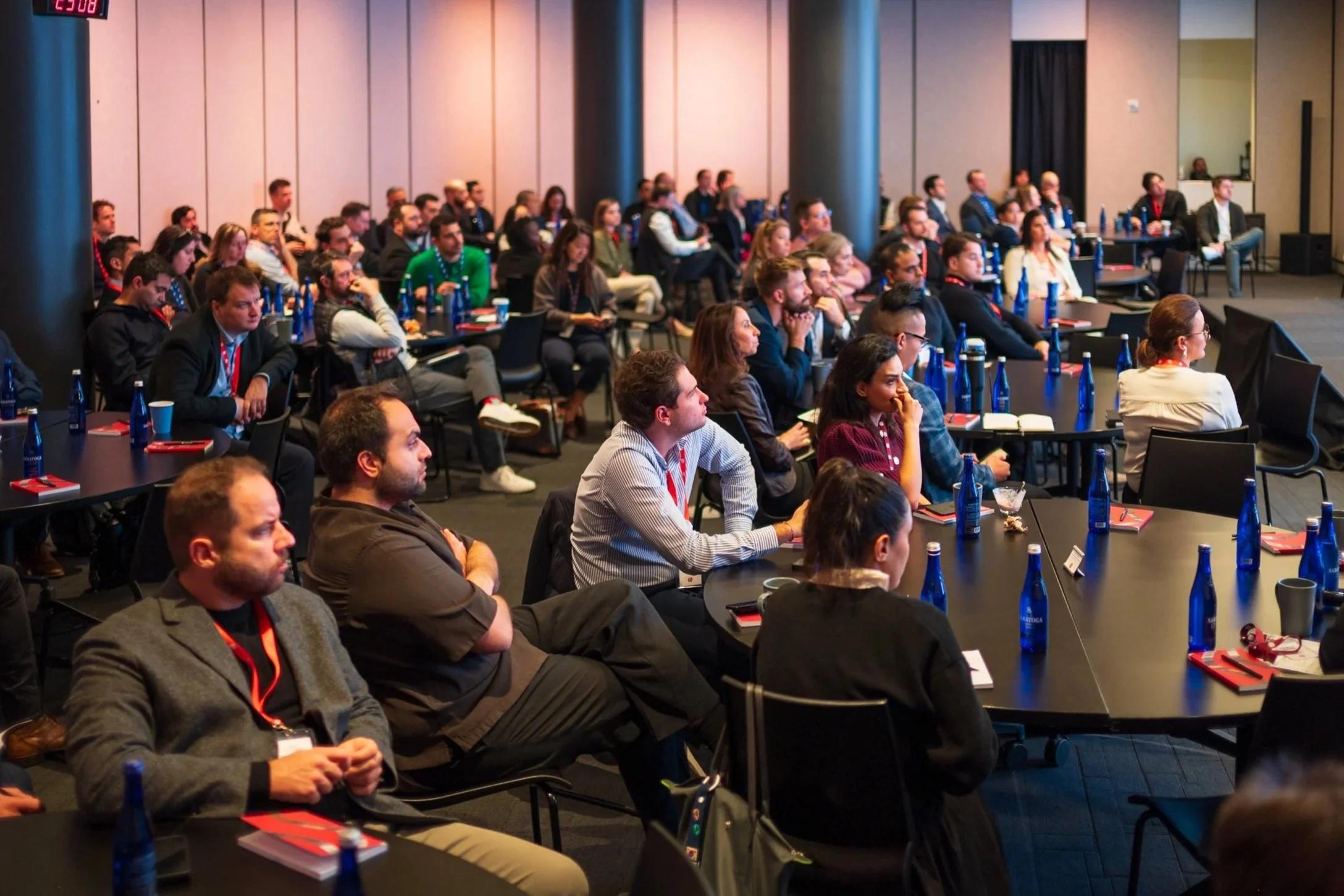 Audience attending a conference or seminar in a large room with a presentation screen at the front, sitting at tables with water bottles and notebooks.