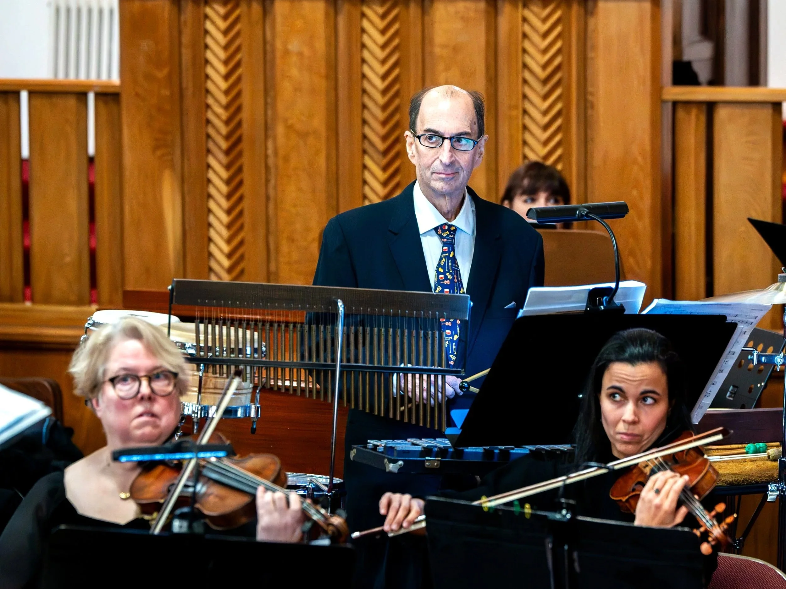 A family orchestra performing on stage, consisting of two violinists, a man in a suit and colorful tie, and a woman in the background.