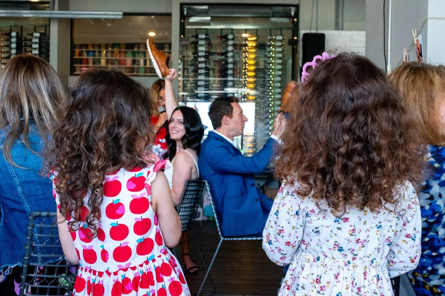 A group of people at a party or celebration, some sitting and some standing, with women in floral and patterned dresses, and children with curly hair in colorful dresses, in a decorated indoor venue.