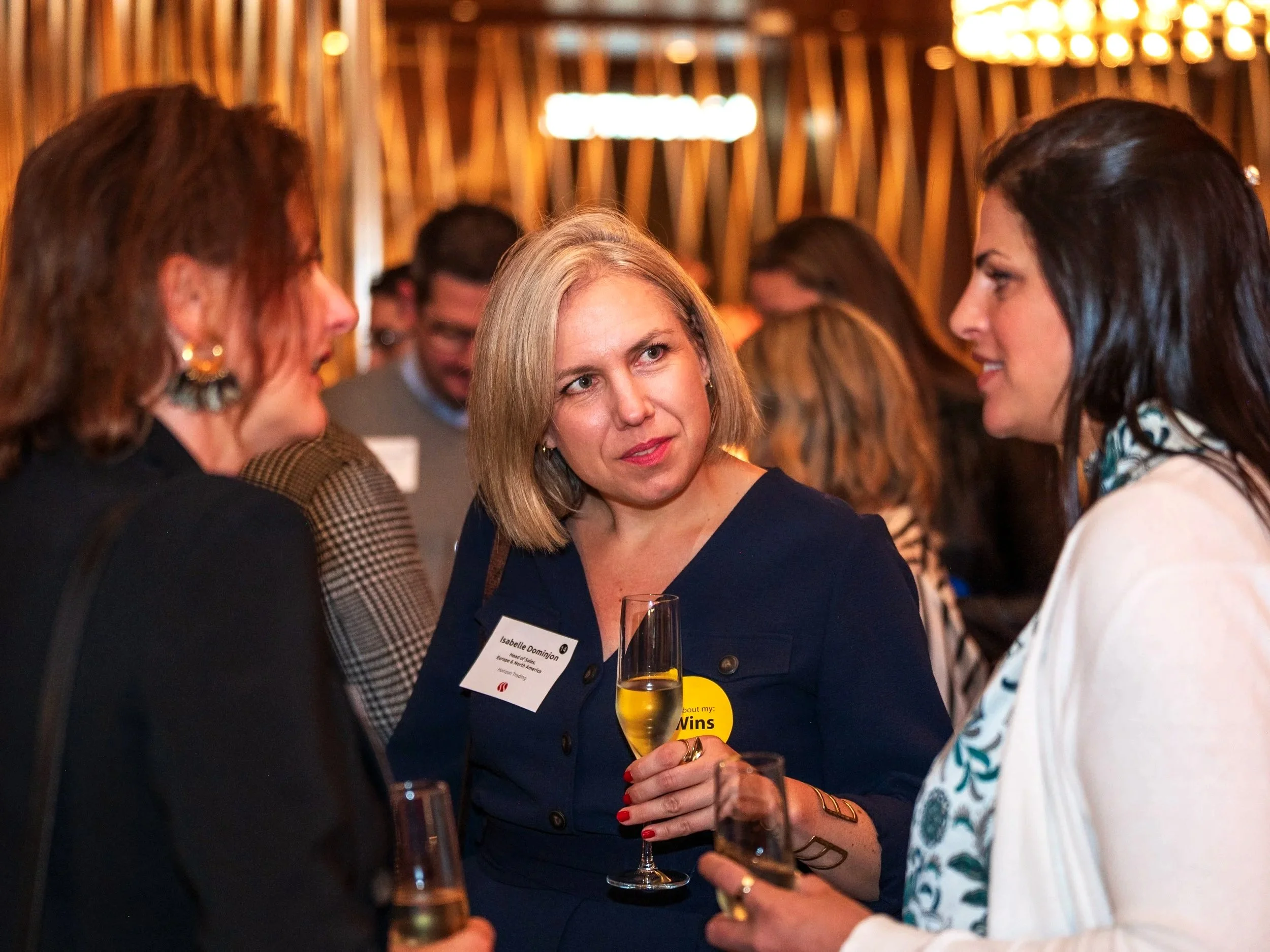 Women at a networking event conversing, some holding wine glasses, with warm lighting and wooden decor in the background.