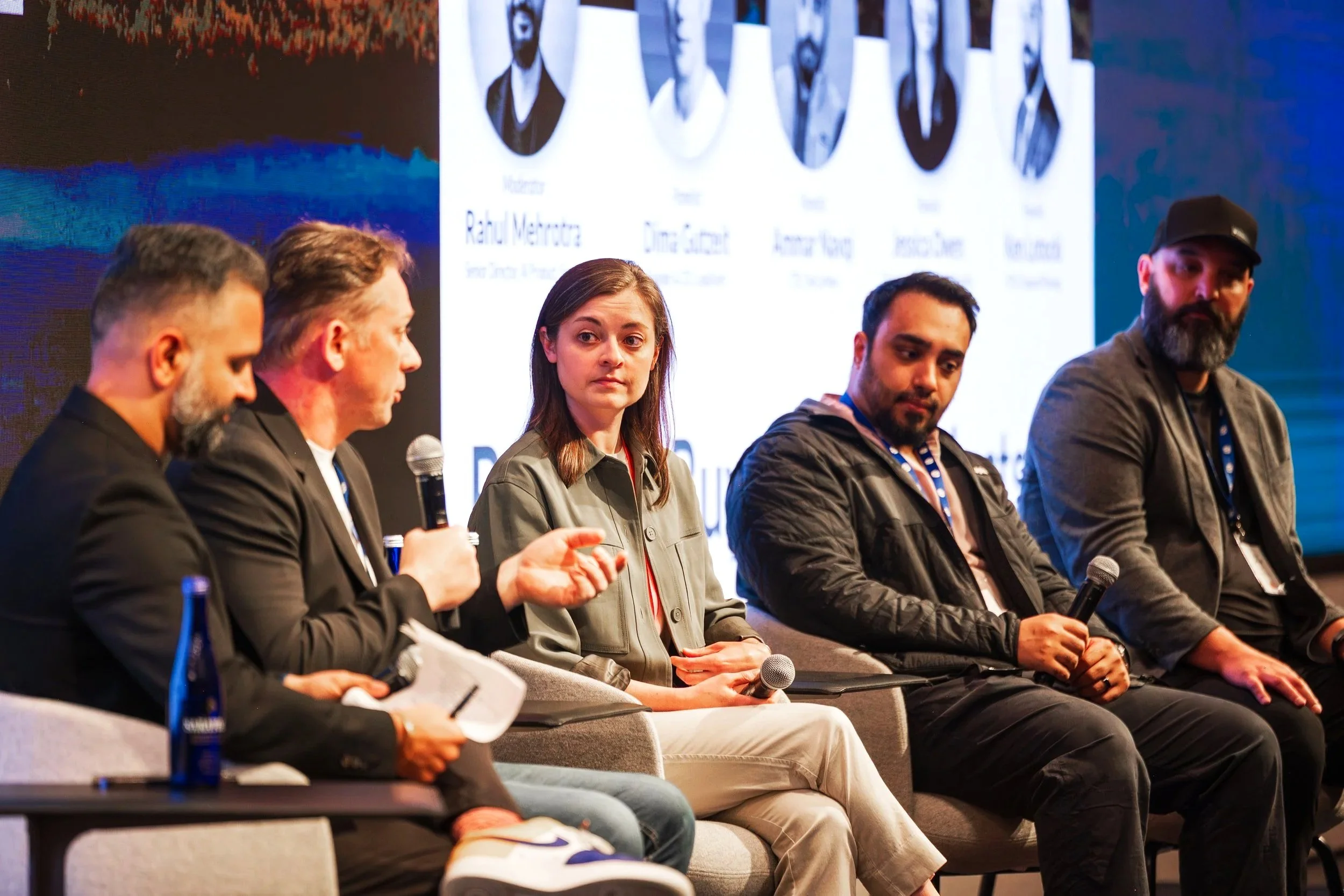 Five people sitting on chairs on a stage during a panel discussion, with a large screen displaying the panelists' photos and names behind them.