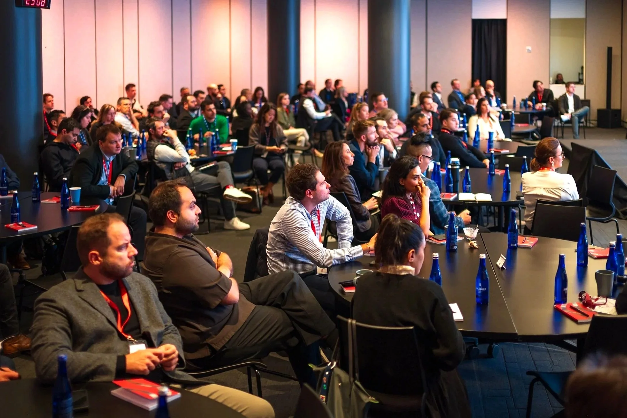 Audience attending a conference or seminar in a large room with a presentation screen at the front, sitting at tables with water bottles and notebooks.